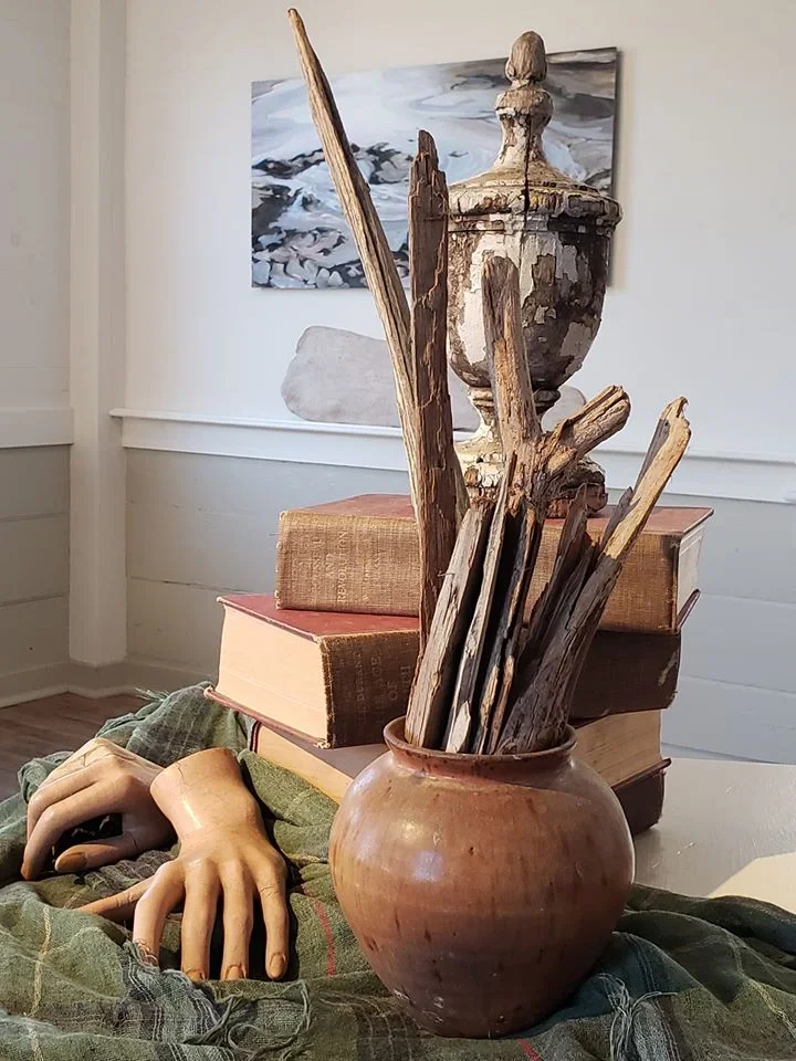 Ceramic vase filled with driftwood sticks, stacked books, a wooden mannequin hand, and a painter's palette arranged on a table with a green cloth, with a painting hanging on the wall in the background.