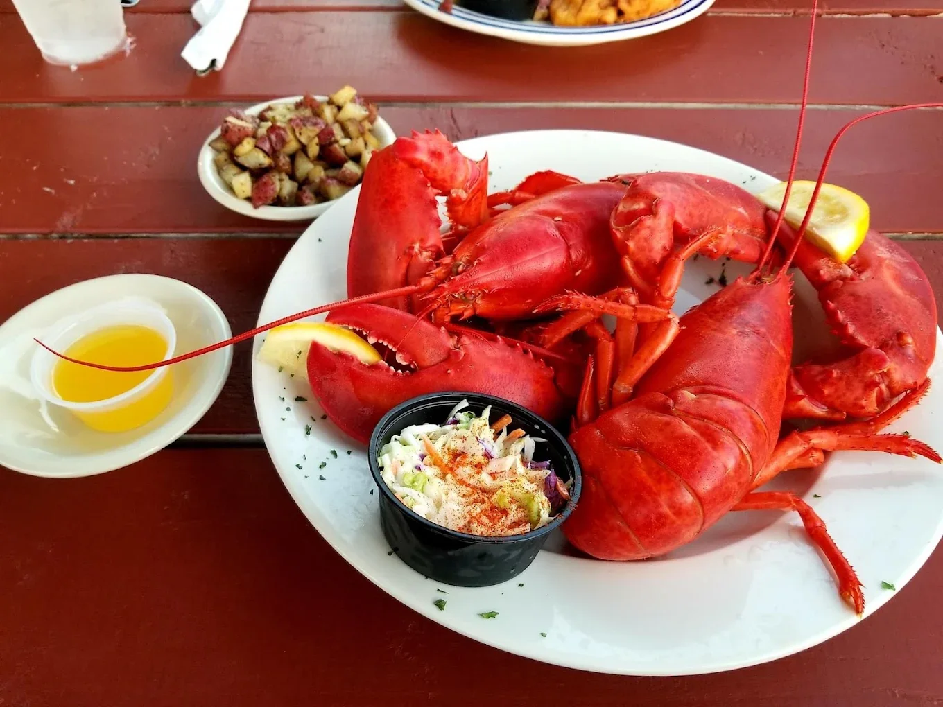 A plate of boiled lobsters with lemon wedges, a side of melted butter, and a small bowl of coleslaw on a wooden table.