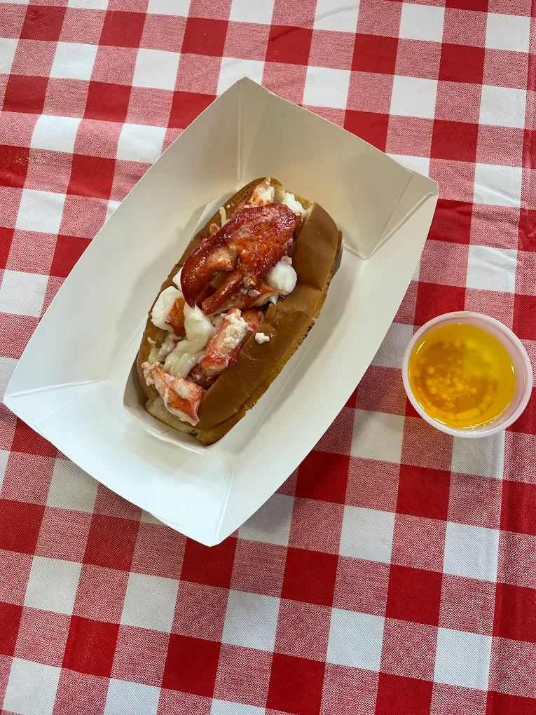 Lobster roll with lobster meat in a toasted bun inside a white paper tray, accompanied by a cup of melted butter, all on a red and white checkered tablecloth.