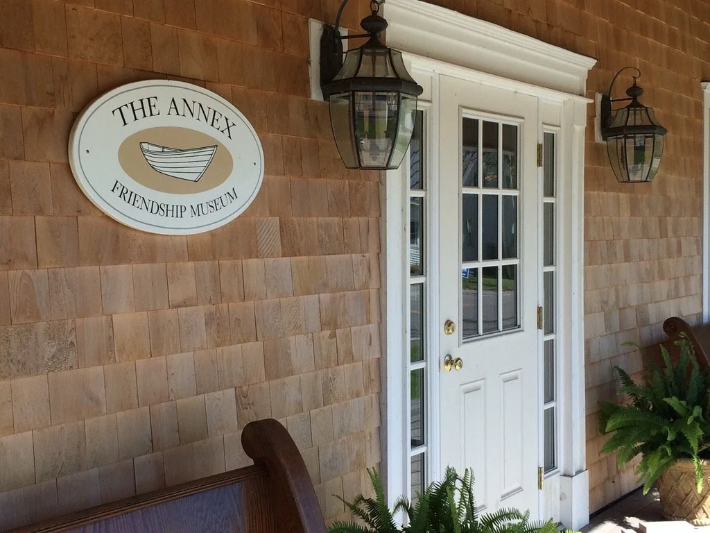 Exterior of a building with a sign that reads "The Annex Friendship Museum," showing a boat illustration, and a white door with glass panes, flanked by two lantern-style outdoor wall lights and potted plants on either side.