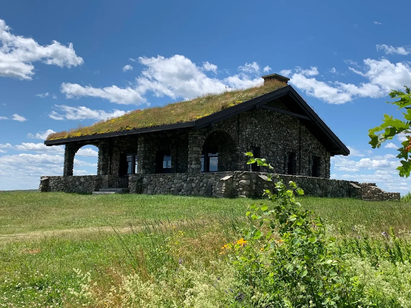 A stone house with a grass-covered roof sits on a grassy hill under a blue sky with scattered clouds.