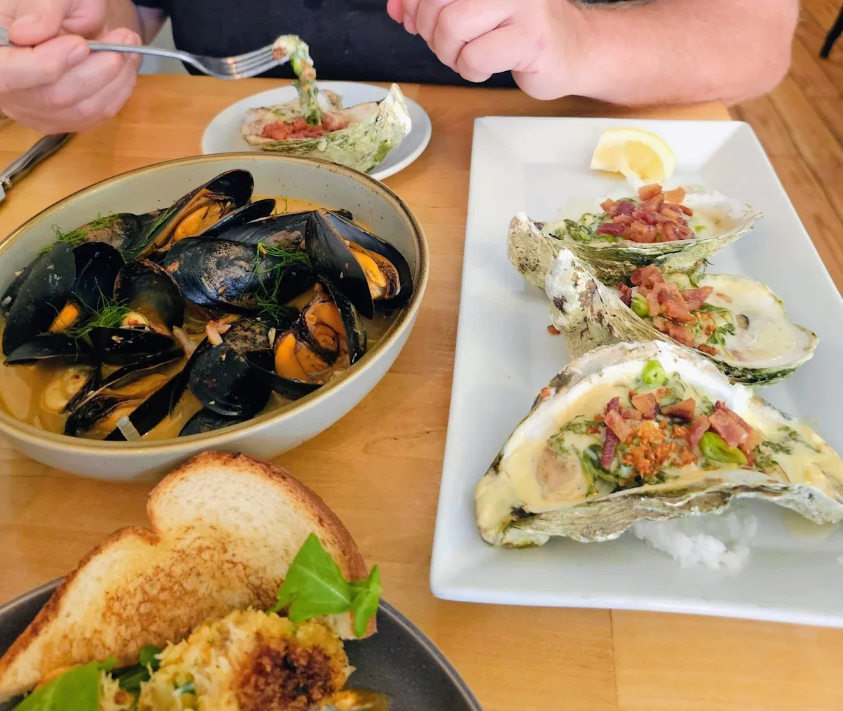 A plate of grilled oysters topped with bacon and green onions, a bowl of steamed mussels in broth, and a plate of toasted bread with a green leaf garnish.