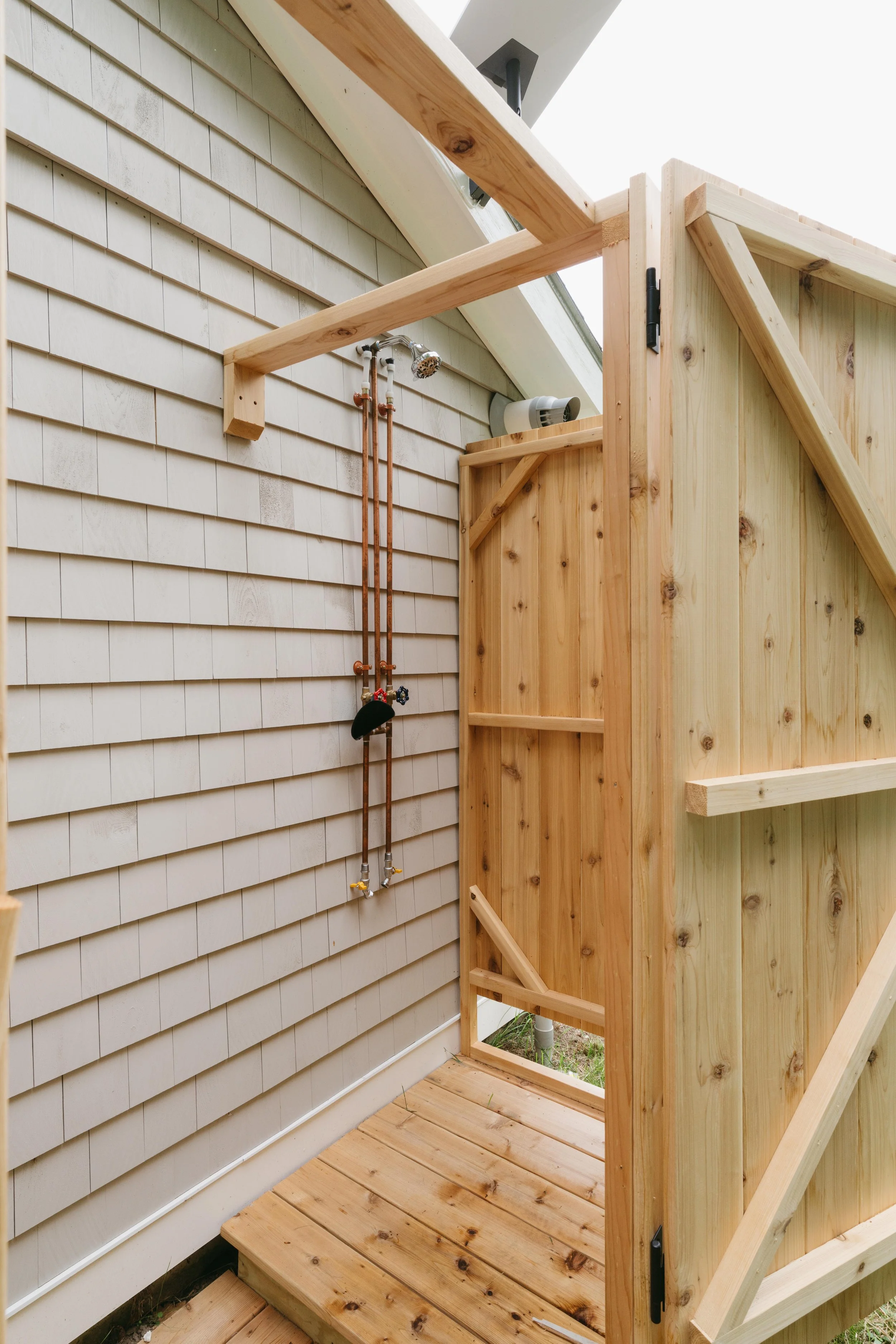 The image shows an outdoor shower area under construction, with a wooden frame and a small wooden platform. There are exposed plumbing pipes with a showerhead attached to the wall, and a white vent pipe above.