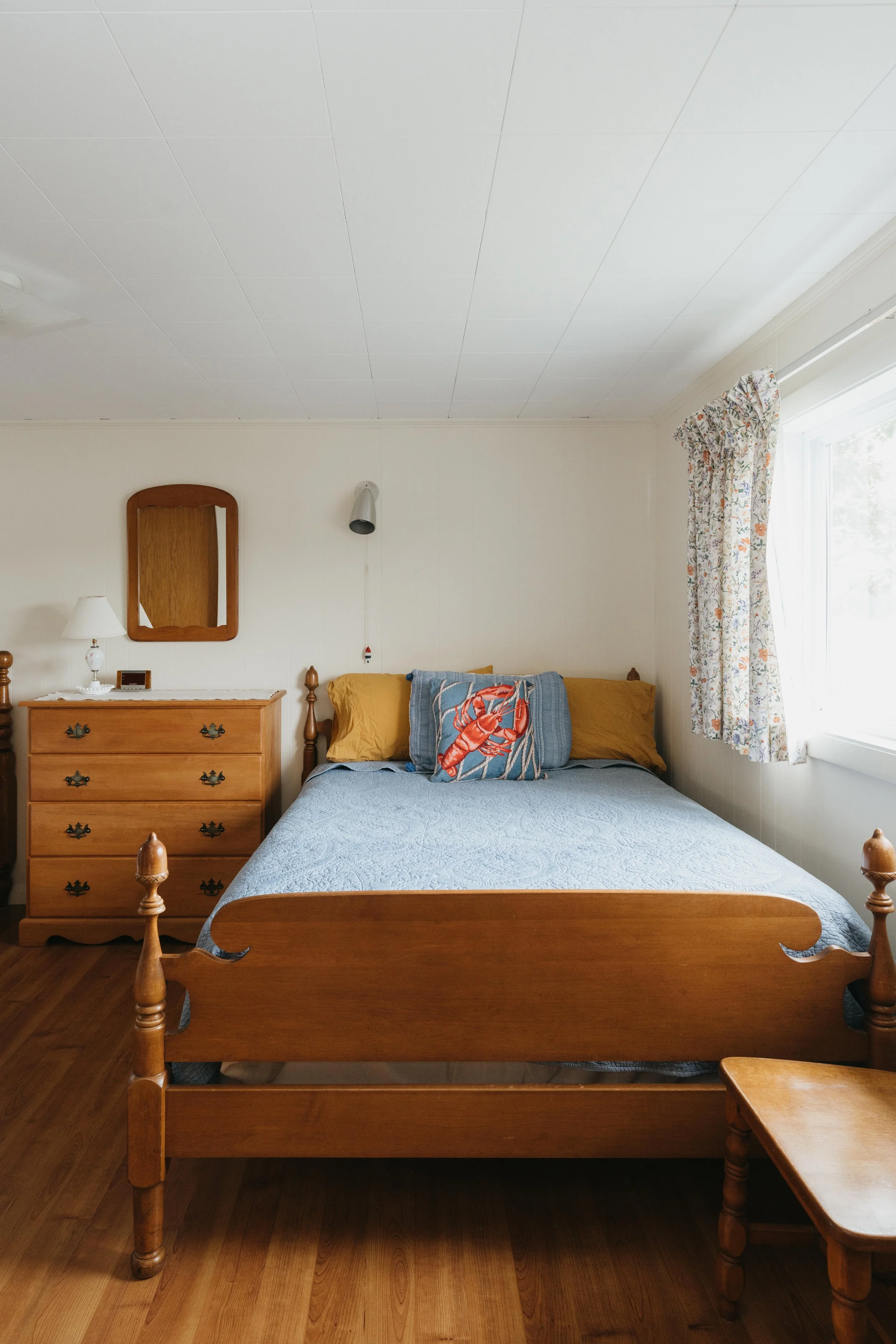 A bedroom with a wooden bed frame, a blue quilt, yellow pillows, a decorative travel pillow, a wooden dresser with a mirror, a lamp, a window with floral curtains, and hardwood flooring.