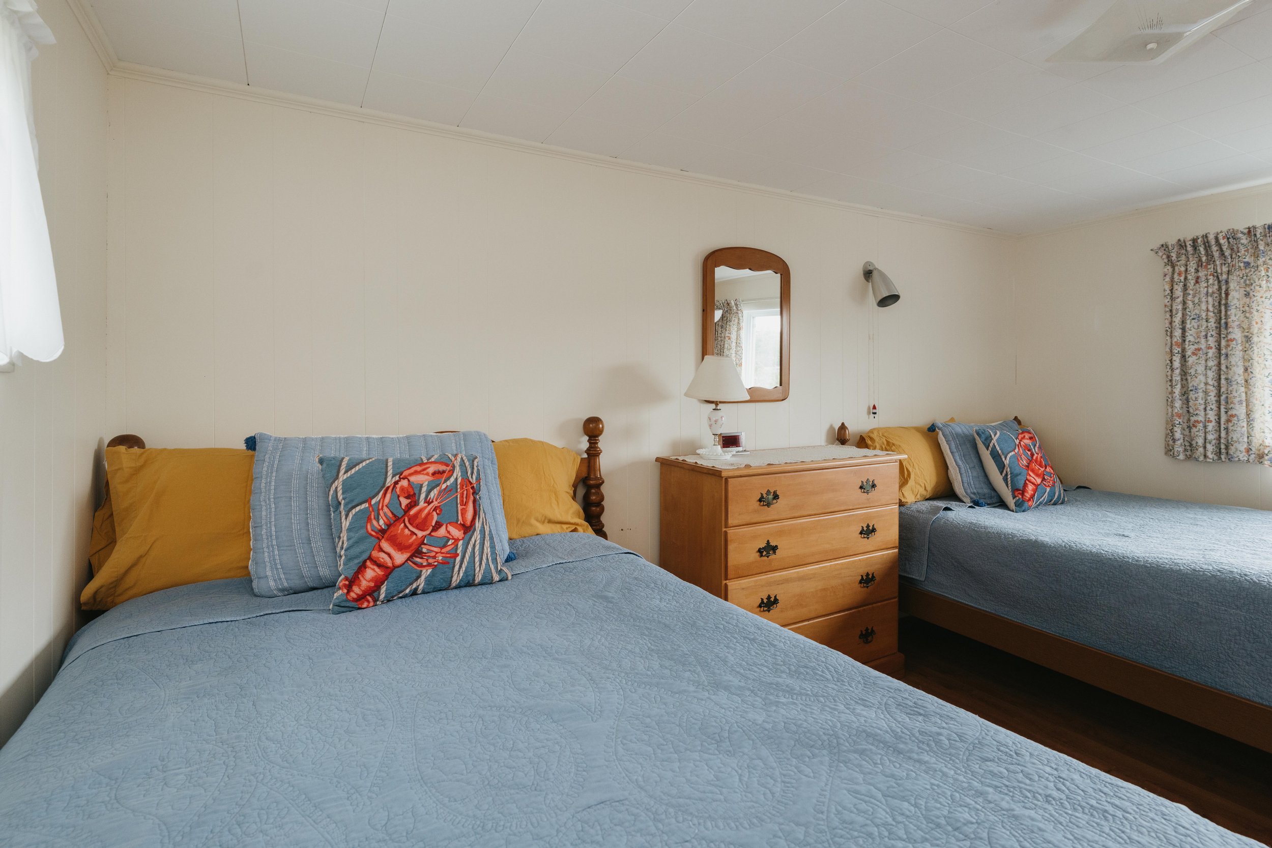 Two beds with blue bedspreads, one with yellow pillows and a decorative pillow featuring a lobster, the other with yellow pillows and a similar decorative pillow, a wooden dresser with a lamp and mirror, floral curtains, and hardwood floor.