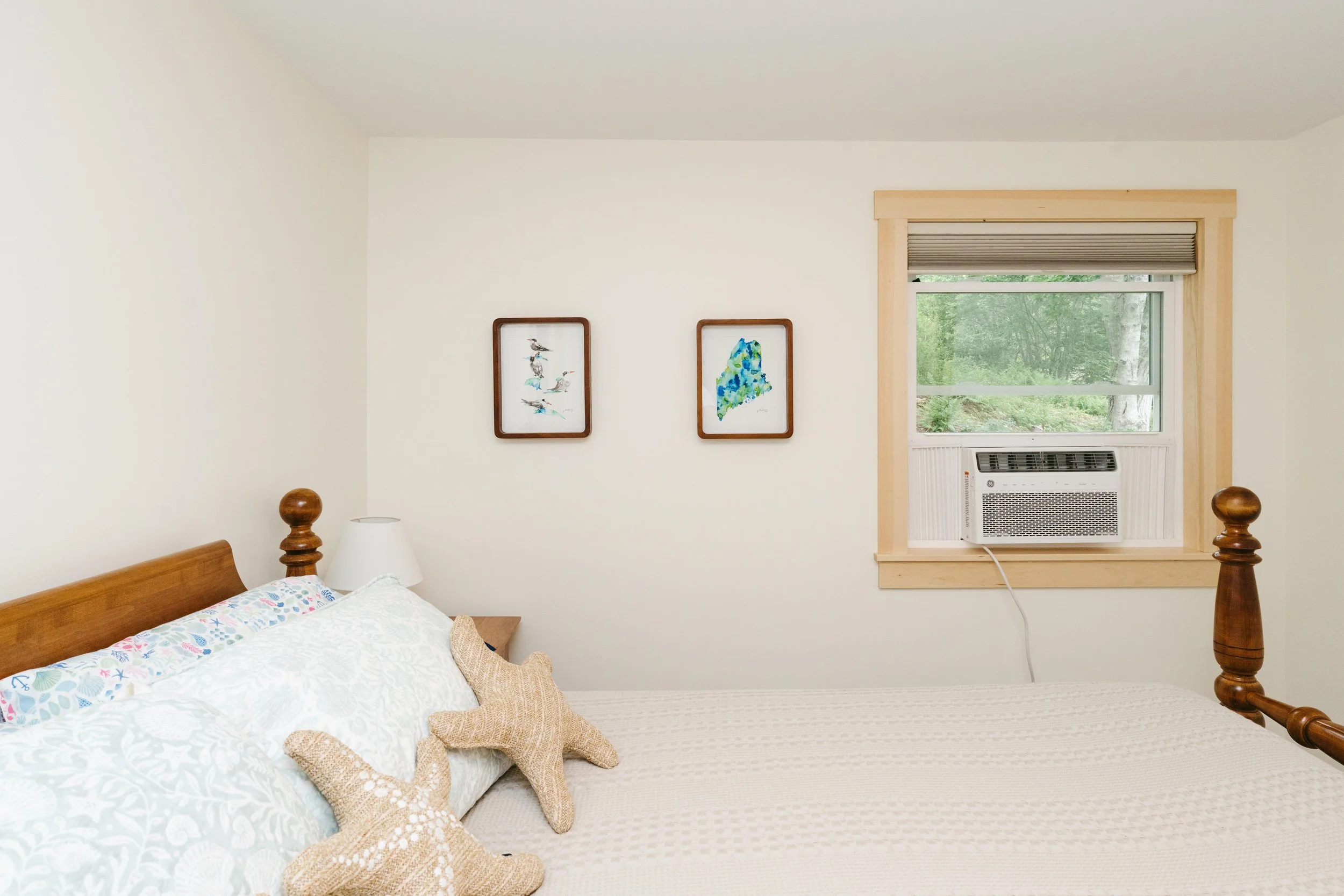 Bedroom with a wooden bed frame, starfish pillows, patterned bedspread, window with a view of greenery, wall art with bird illustrations, air conditioning unit in the window
