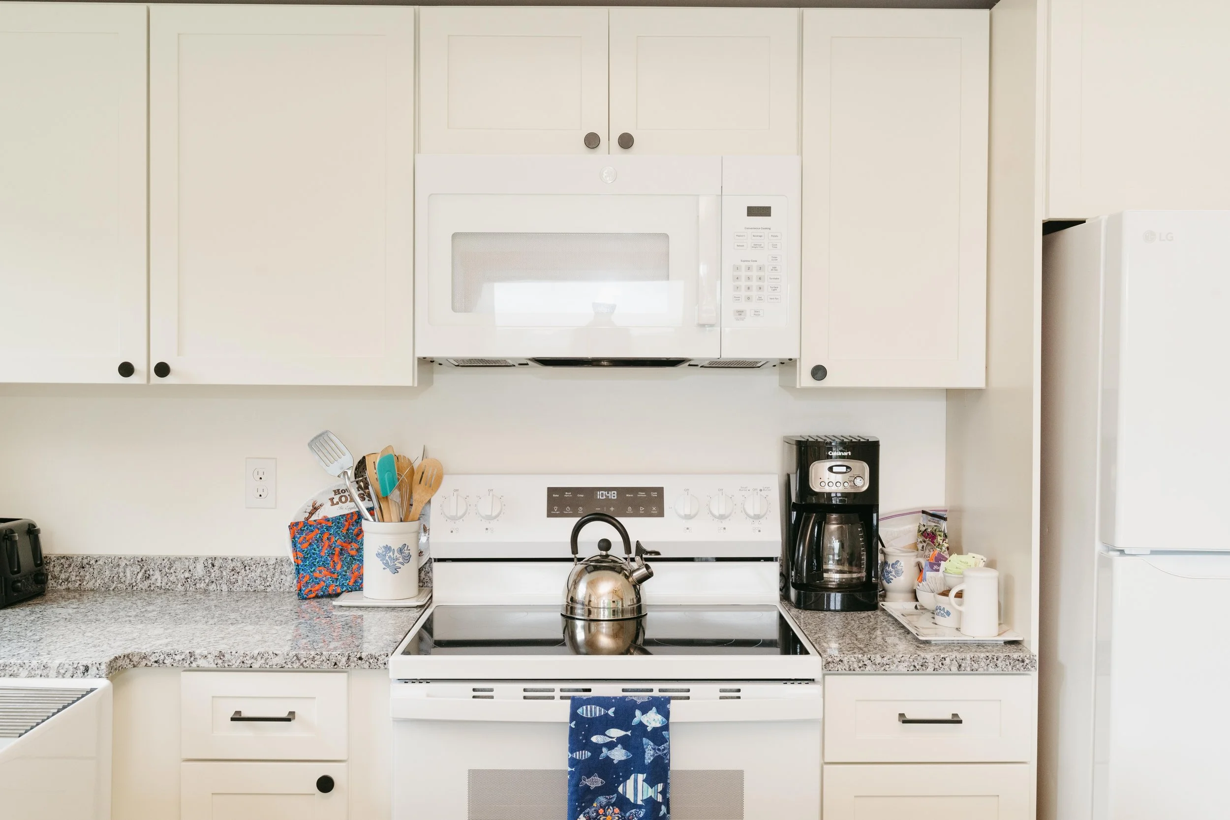 Kitchen with white cabinets, a microwave, coffee maker, kettle, and various utensils and mugs. A stove with a tea towel hanging from it, and a refrigerator on the right.