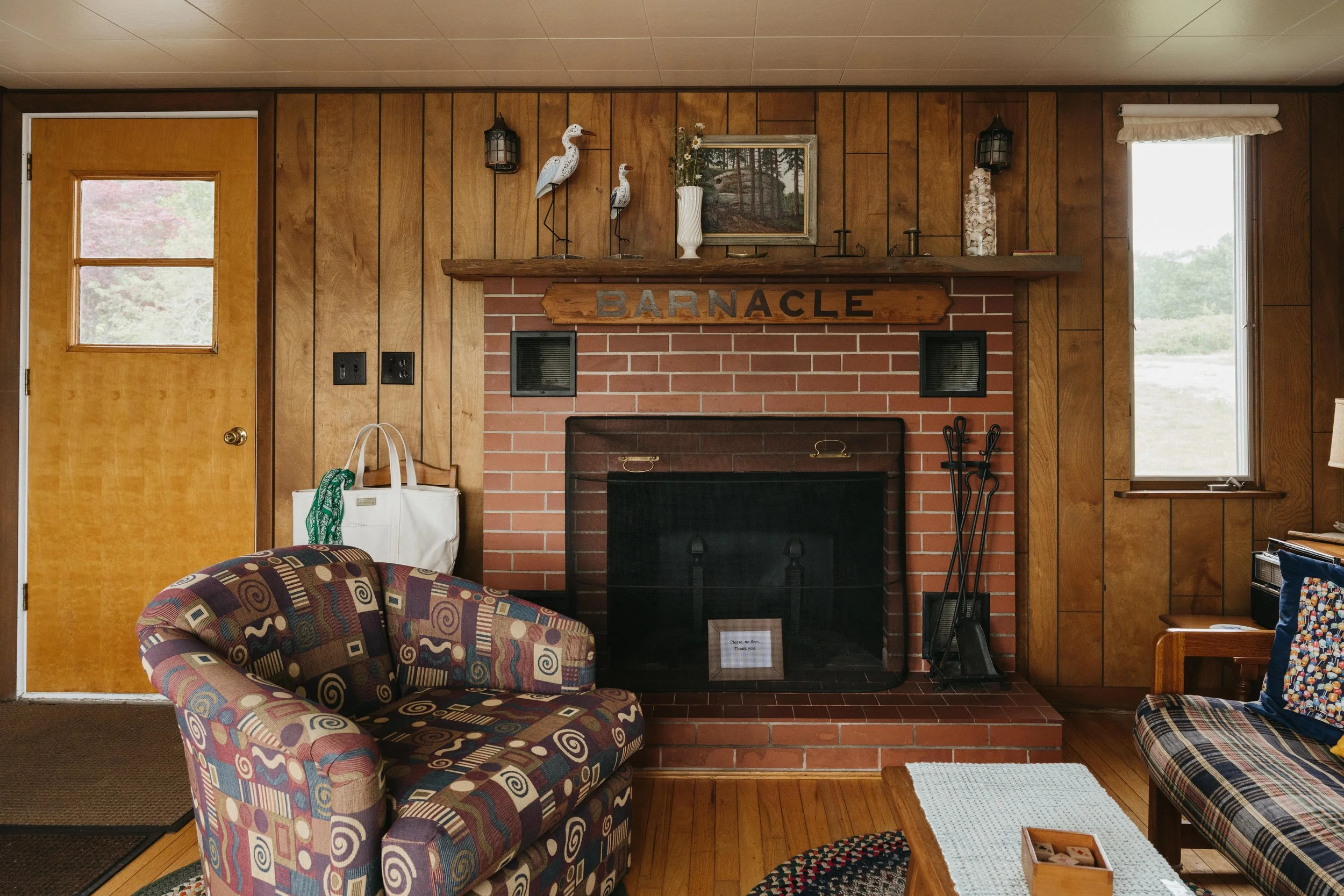 Living room with wood-paneled walls, a brick fireplace, and a wooden mantel with a sign that says "BARNACLE." There are two chairs with patterned upholstery, a small wooden coffee table, a window on the right, and a door on the left. Decorations incl