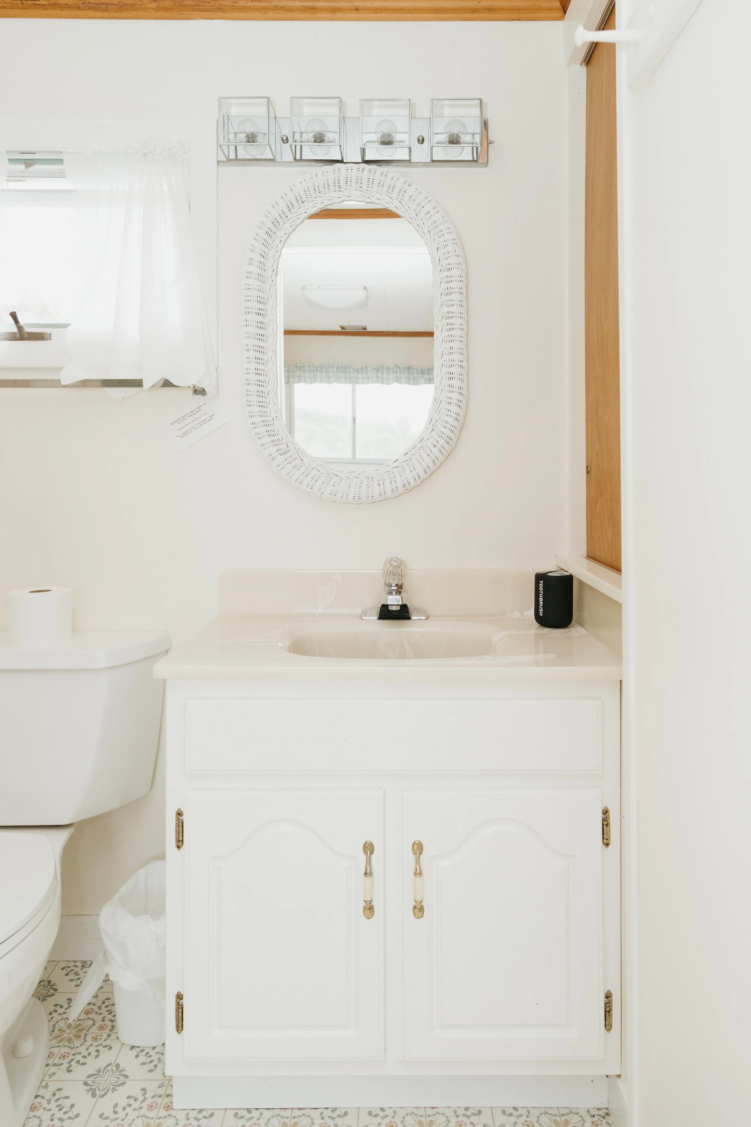 A small bathroom with a white vanity, oval mirror with a white wicker frame, wall-mounted light fixture, toilet, and patterned tile floor.