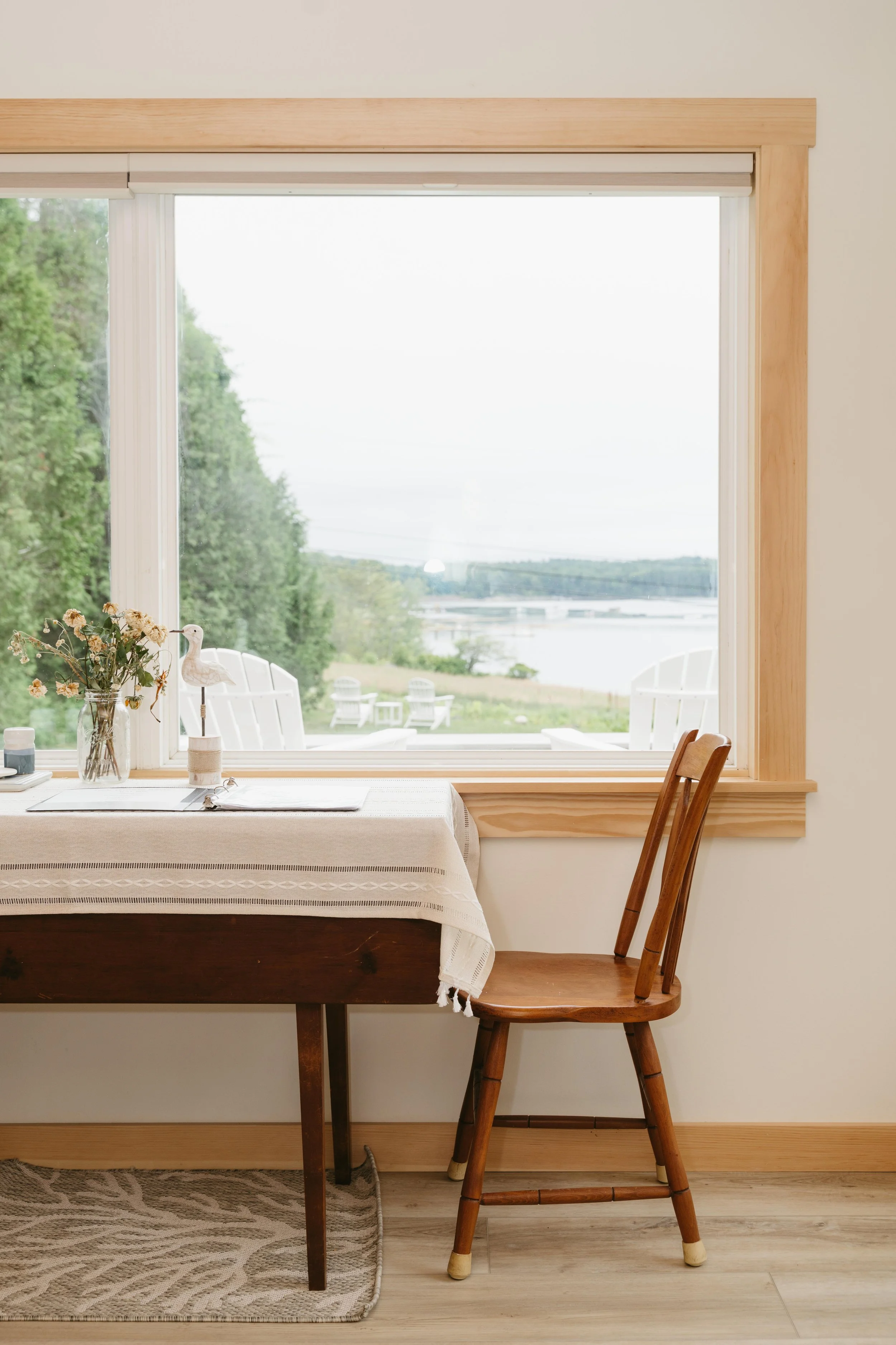 A cozy dining nook with a wooden table covered by a light-colored tablecloth, a wooden chair, a vase with flowers, and a large window overlooking a scenic outdoor view of trees, a body of water, and Adirondack chairs.
