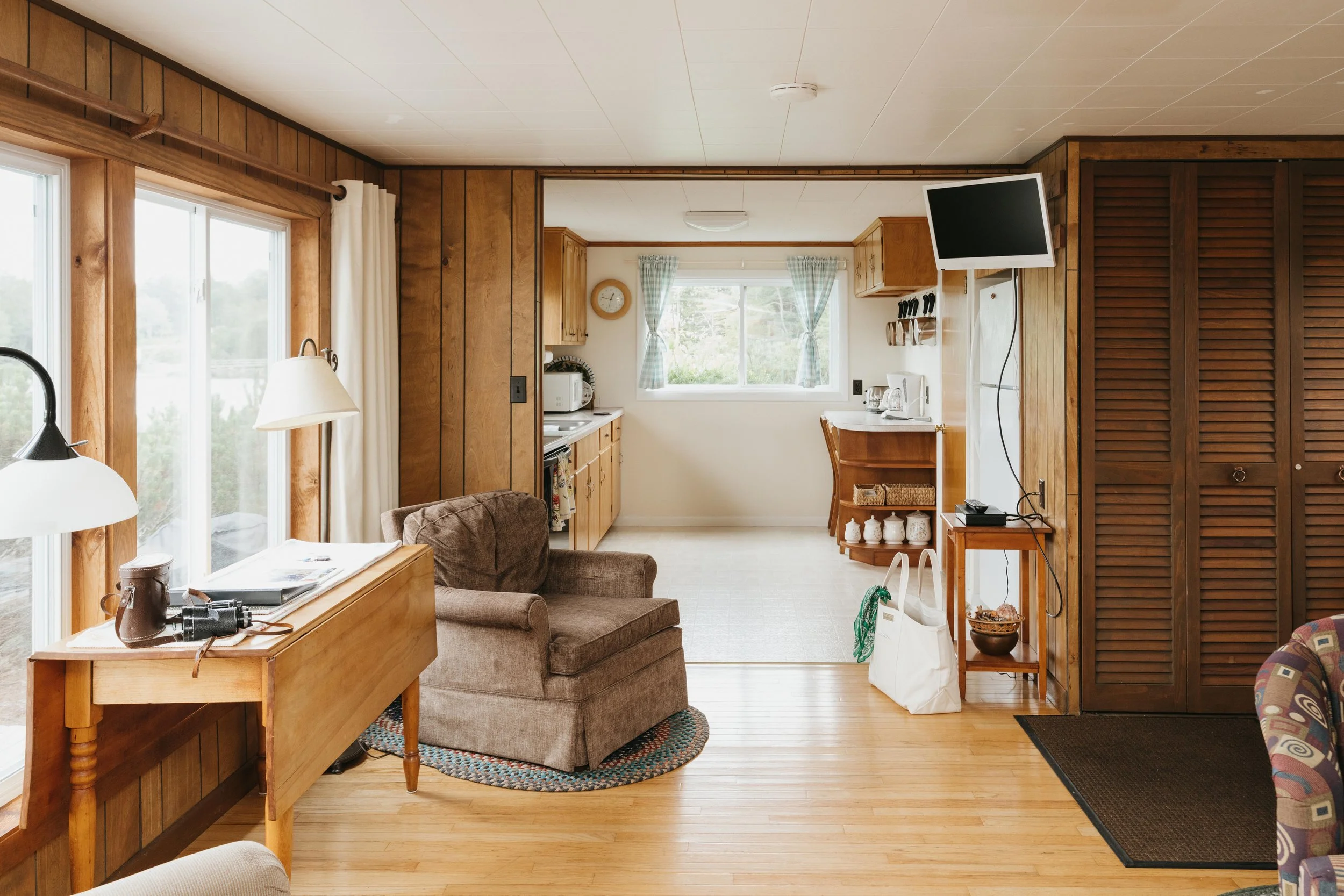 Living room with wood-paneled walls, large windows, and a view of the outdoors; furniture includes a brown armchair, a floor lamp, a small table, and a colorful armchair; kitchen in the background with wooden cabinets, a small desk, and a window with