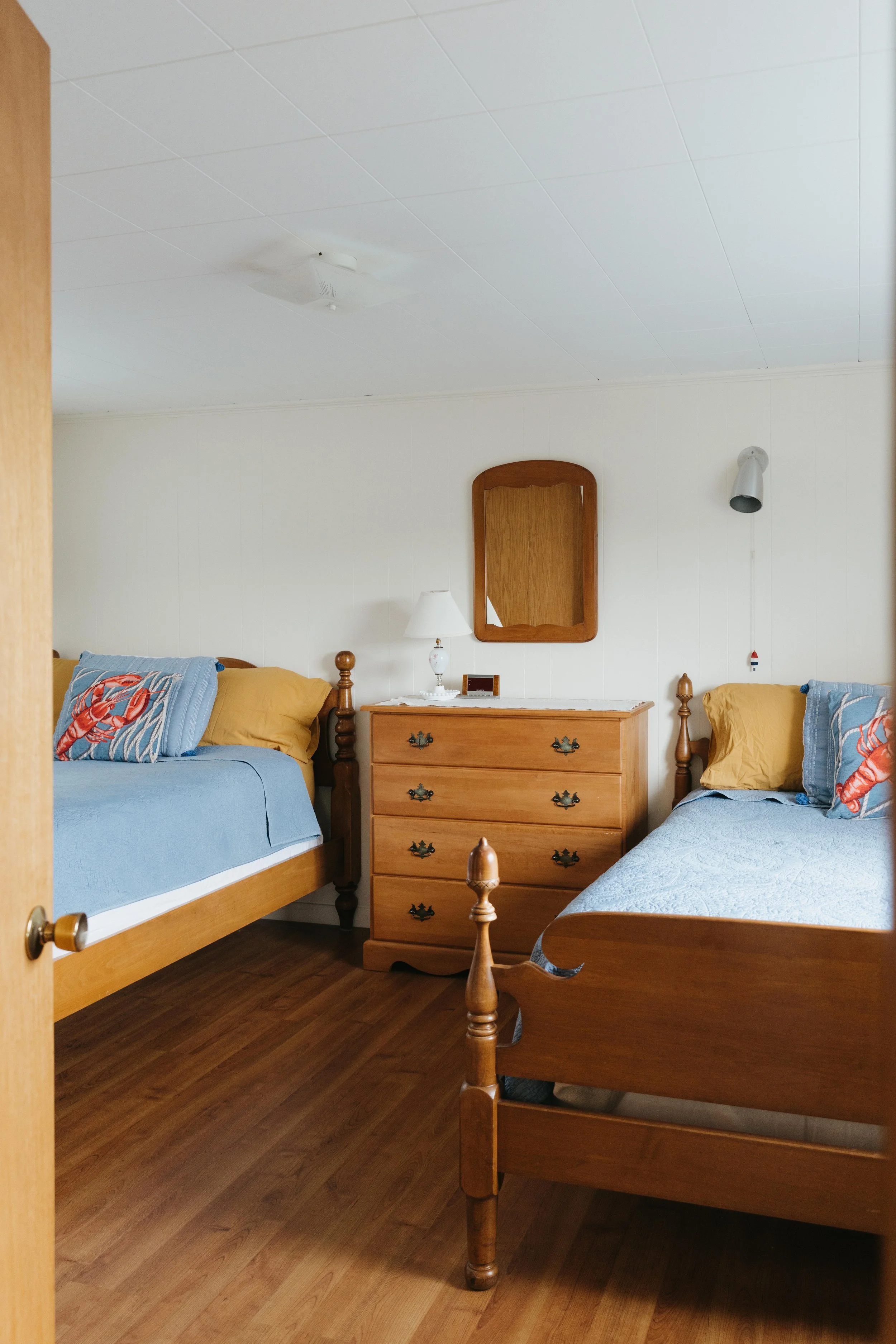 A bedroom with two twin beds with wooden frames and blue bedding, a wooden dresser with a mirror above, a small table lamp, and wall-mounted reading lights, all on a wood floor.