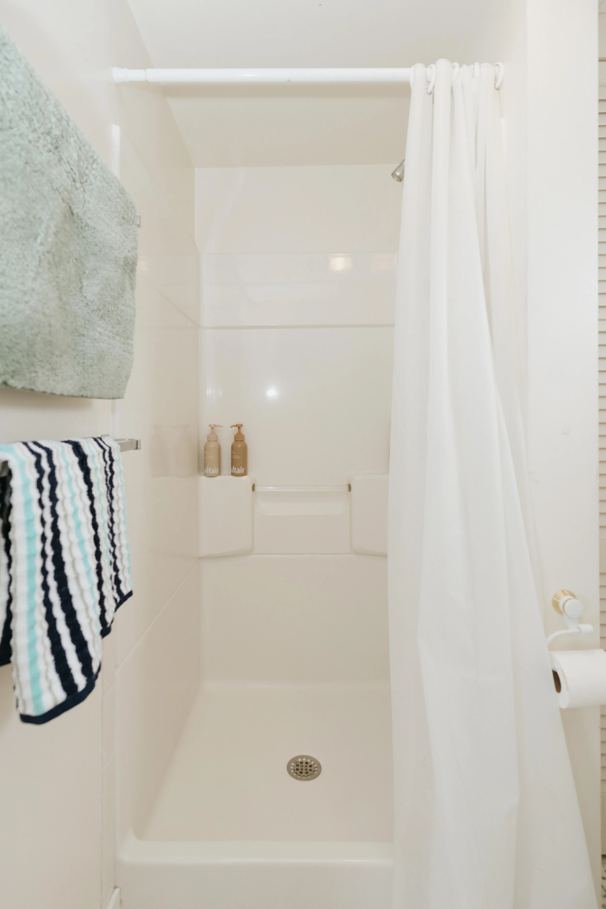 Empty shower with a white curtain, two beige bottles on the built-in shelf, a green towel on the towel bar, and a striped towel hanging, in a small bathroom with white walls.