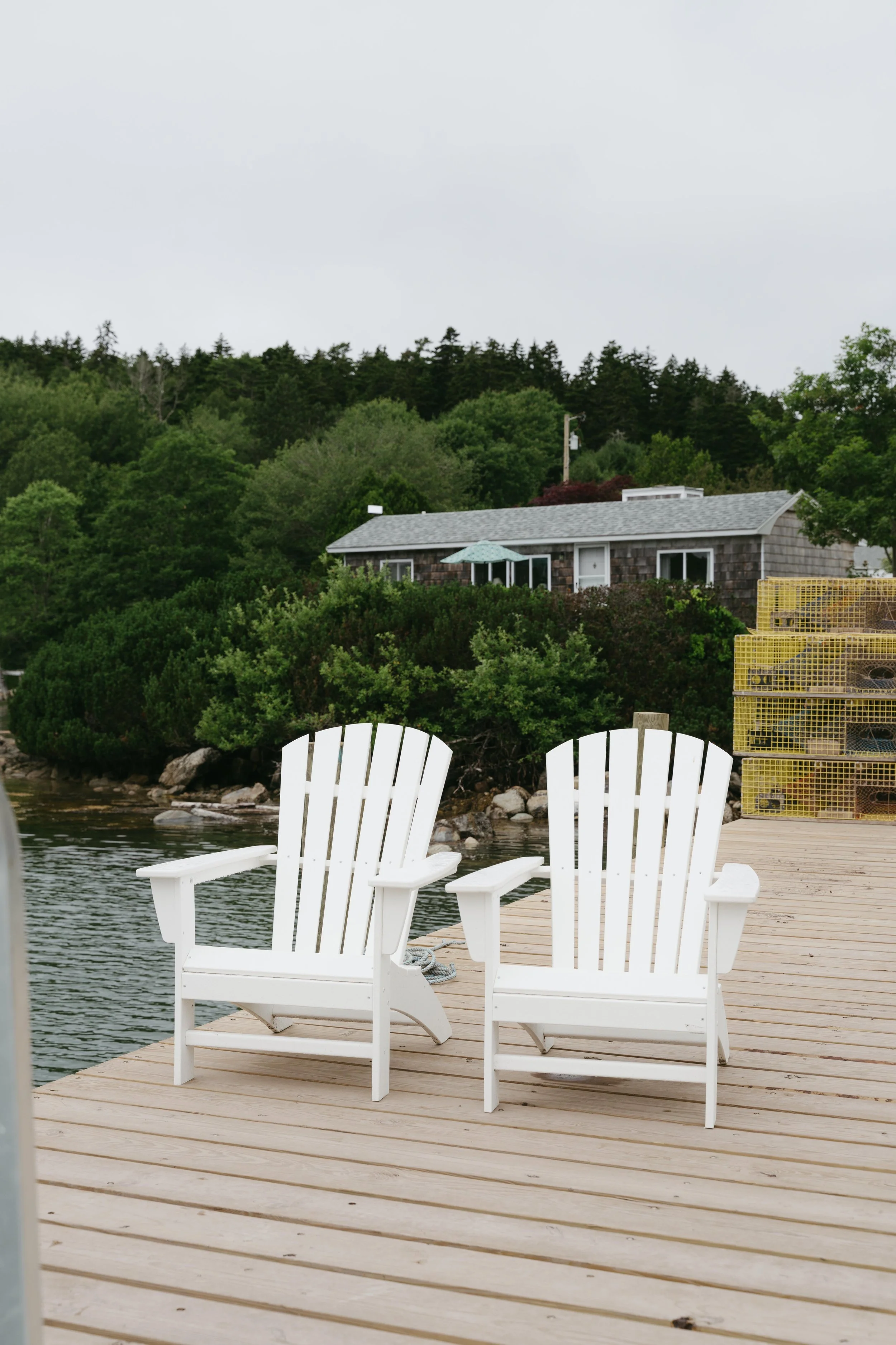 A wooden dock with two white Adirondack chairs facing water, with a house, trees, and a cloudy sky in the background.