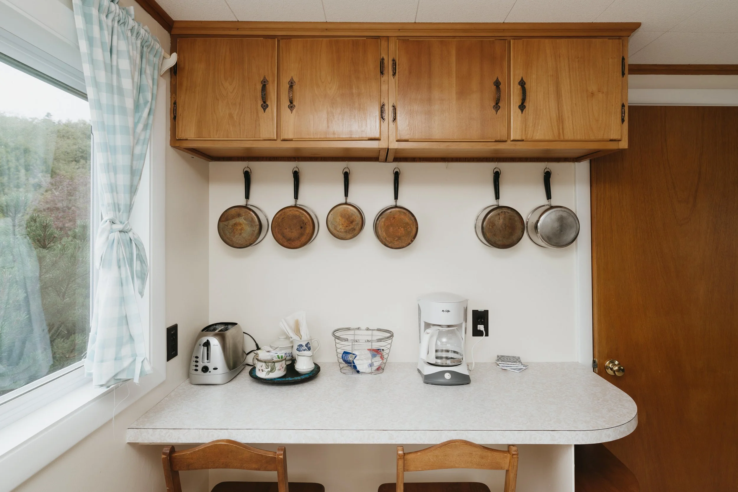 Kitchen counter with coffee maker, toaster, and basket, with six rusty pans hanging on the wall above, and window with checkered curtains on the left.