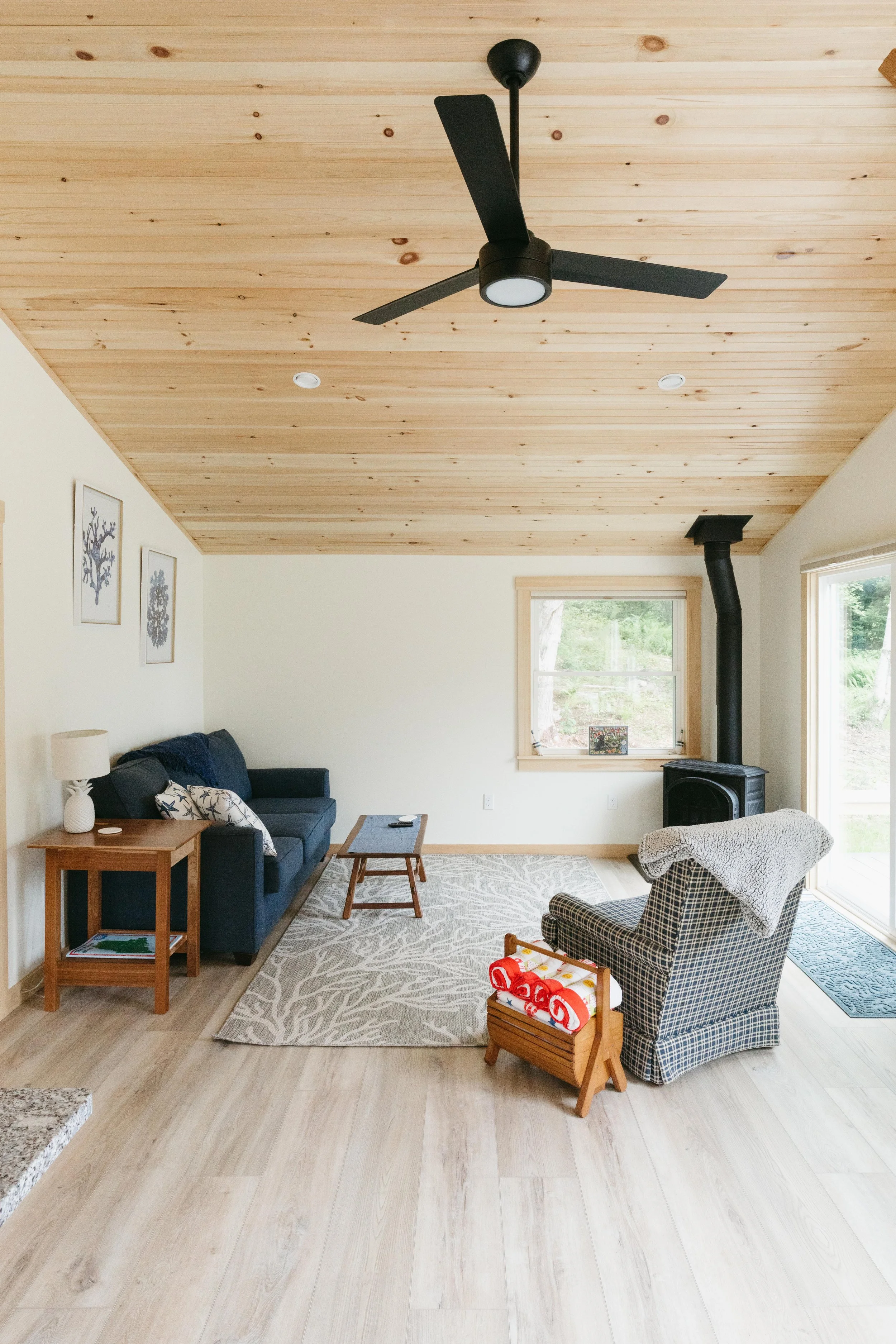 Living room with wood ceiling, ceiling fan, black wood stove, window, sliding glass door, navy sofa, patterned armchair, side table, and area rug.