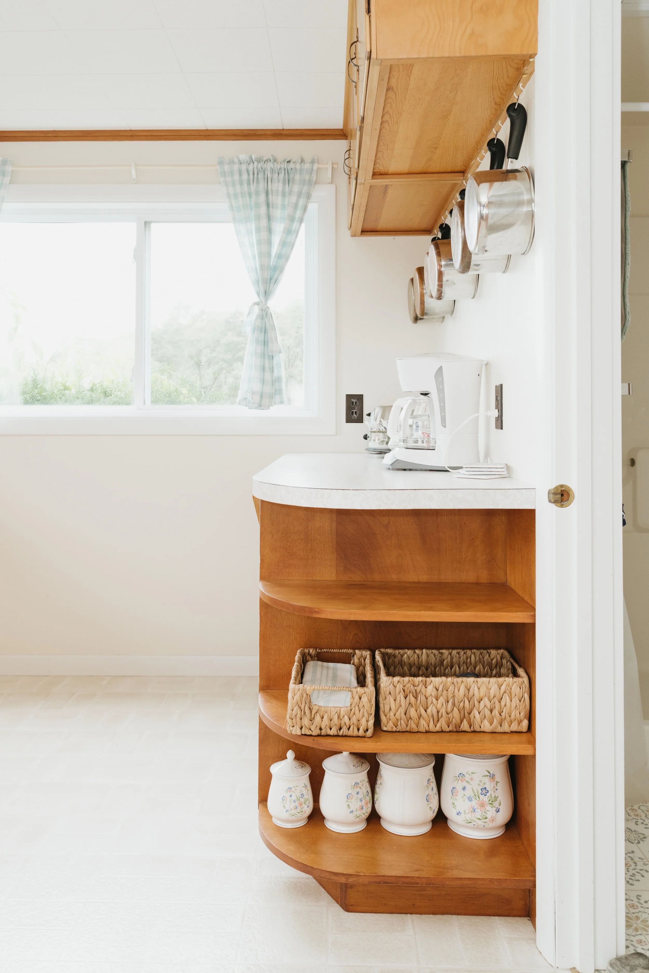 A corner of a kitchen with a wooden shelf holding ceramic jars, a white coffee maker, and woven baskets, next to a window with plaid curtains.
