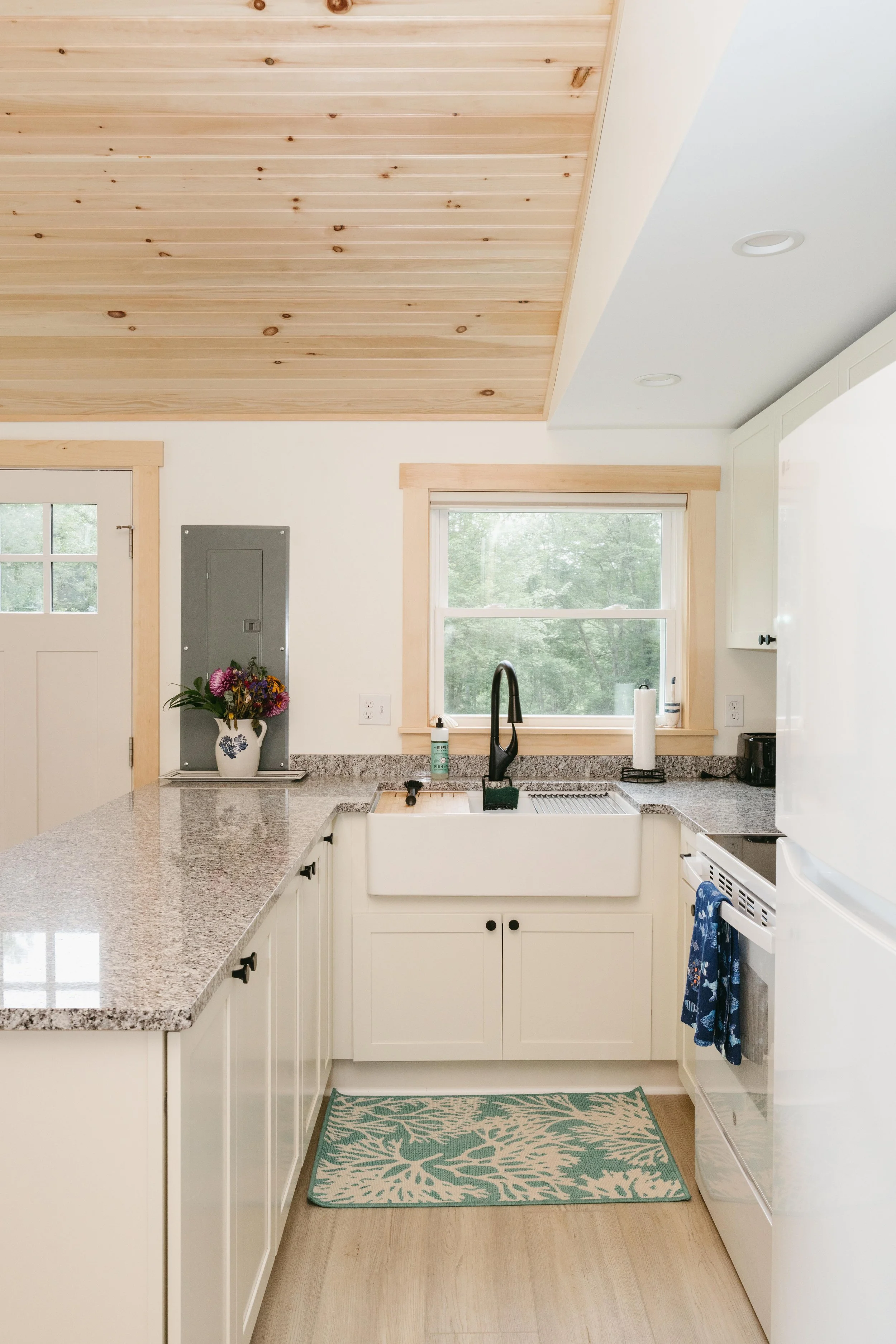 Kitchen with white cabinets, granite countertop, farmhouse sink, window overlooking greenery, and wooden ceiling.