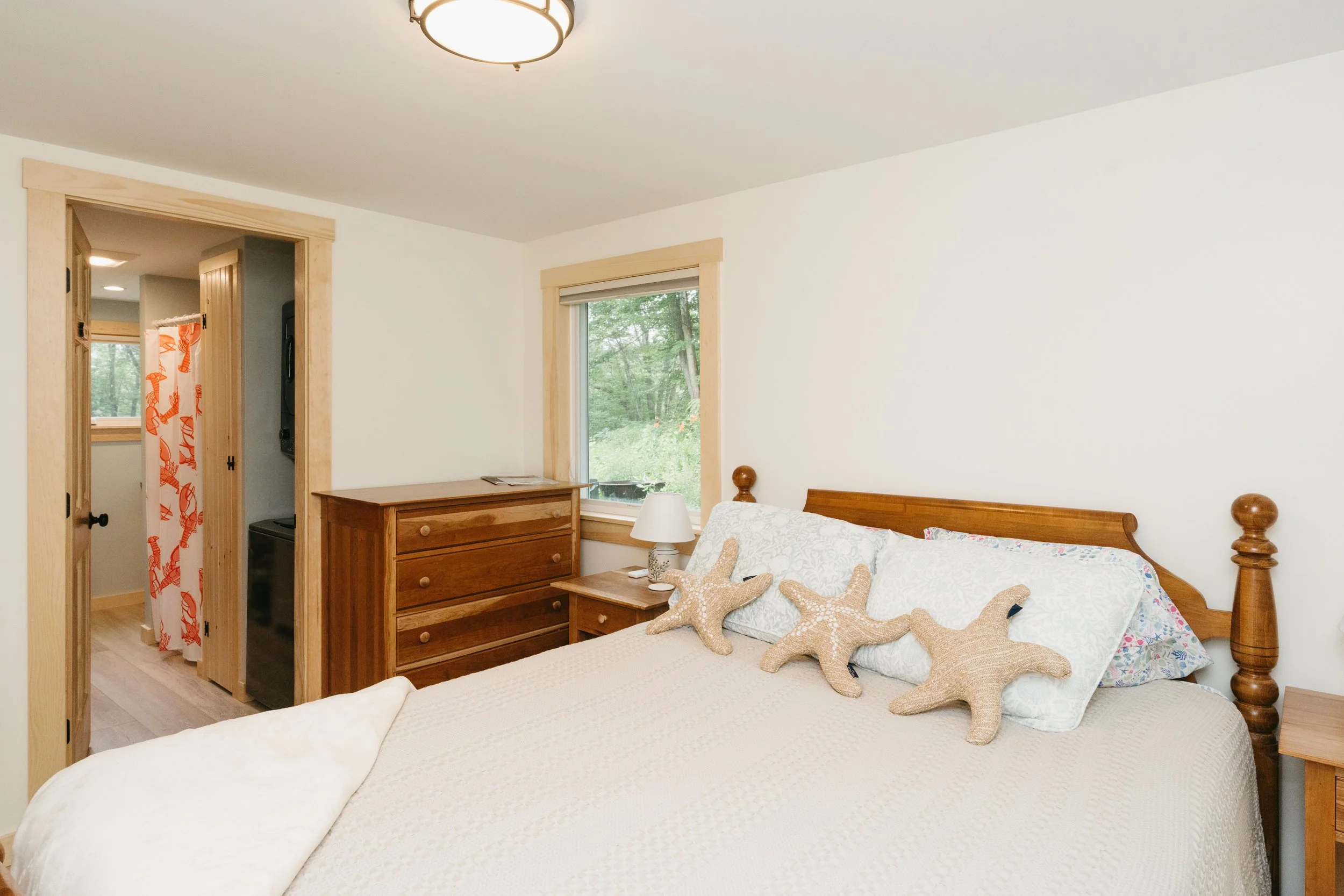 A bedroom with a wooden bed frame, white bedding, and decorative starfish pillows. A nightstand with a lamp is beside the bed. There is a dresser near a window with a view of greenery outside. A small room with a curtain featuring orange lobster desi