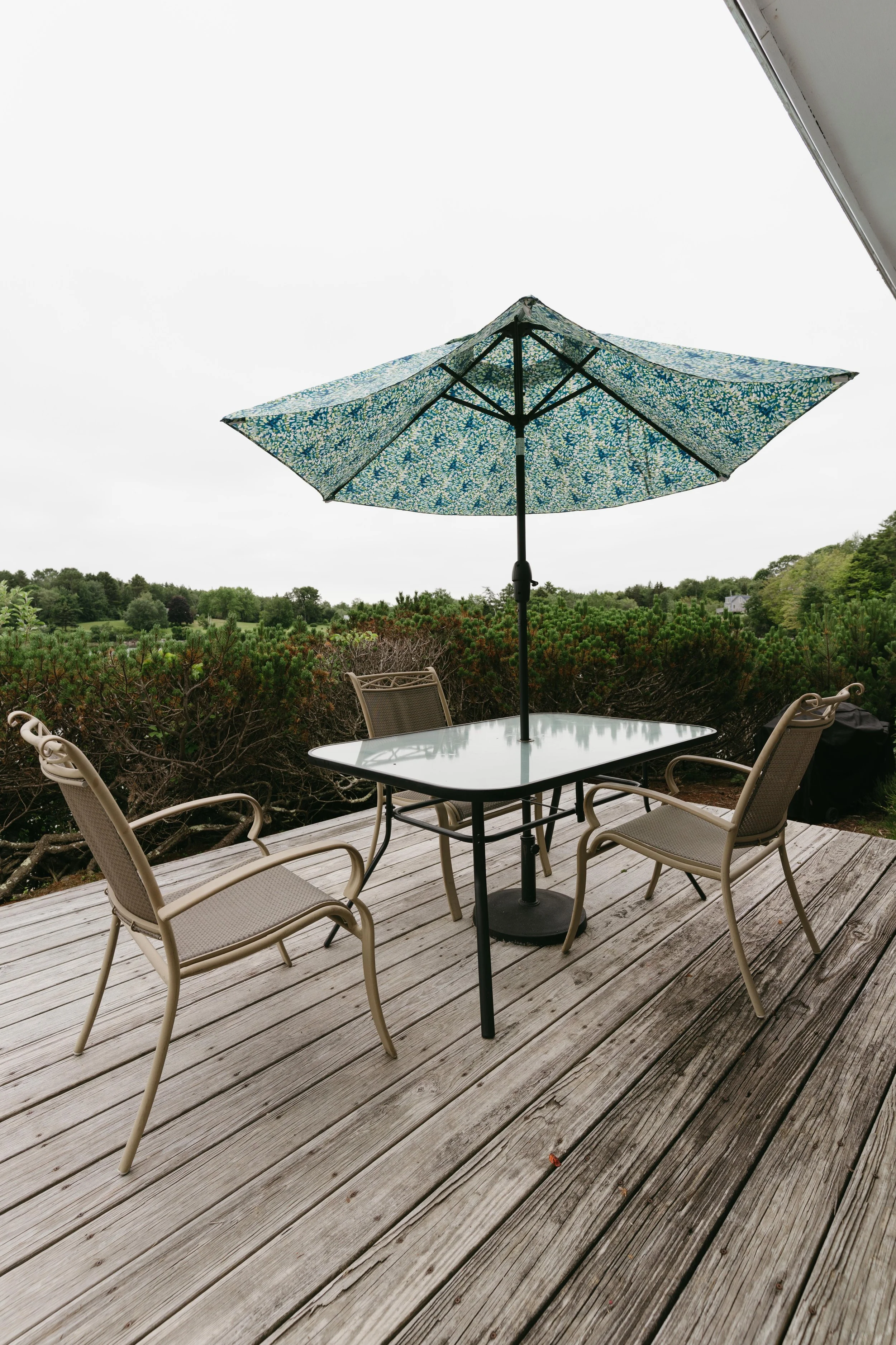 Outdoor patio scene with a glass table, three beige chairs, and a blue patterned umbrella on a wooden deck, overlooking greenery and trees.