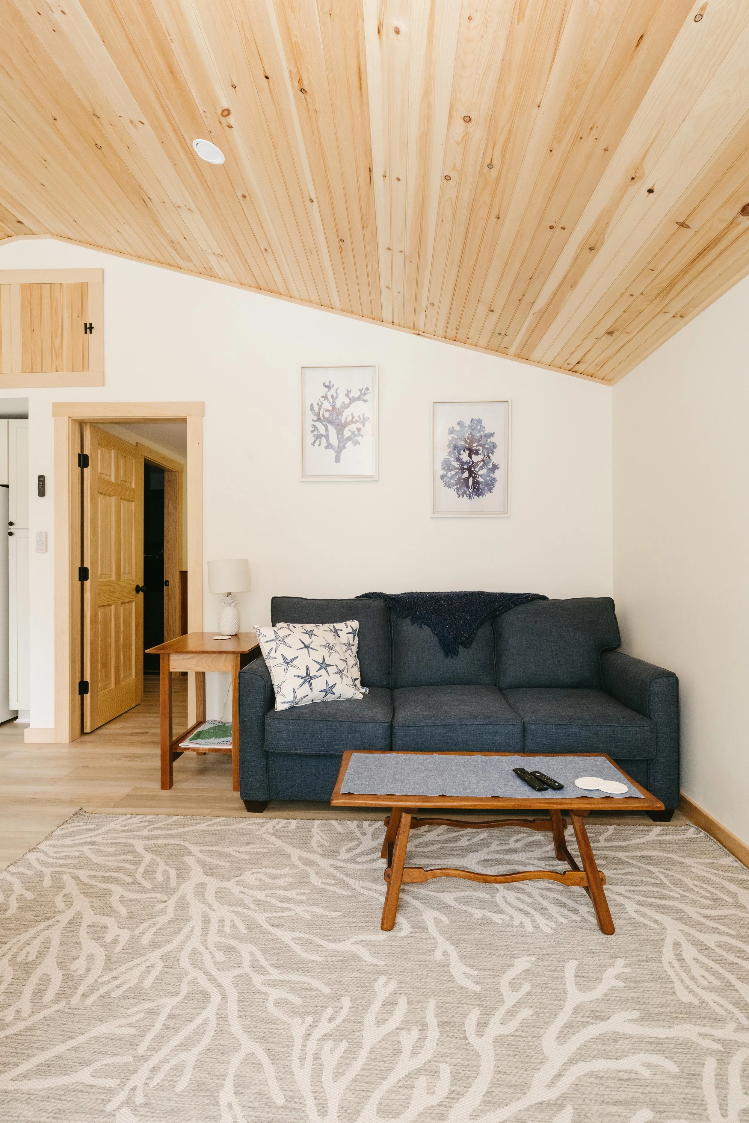 Living room with a dark gray couch, framed artwork of botanical images above, a wooden coffee table with remote controls, a beige patterned rug, a side table with a lamp, and a wooden vaulted ceiling and doorframes.