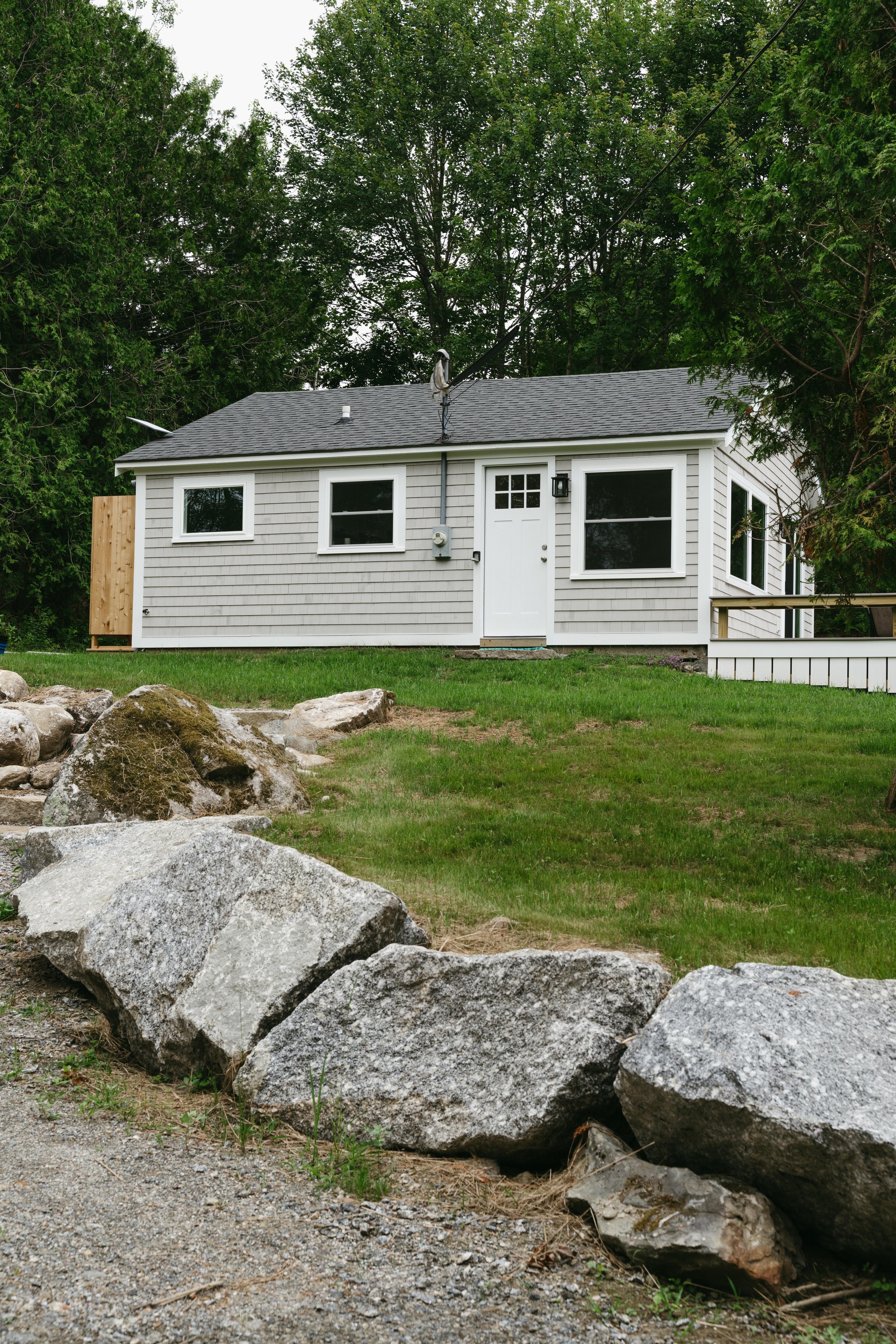 A small house with gray shingles, white trim, and a gray roof, surrounded by green trees and grass, with a rock border in the foreground.