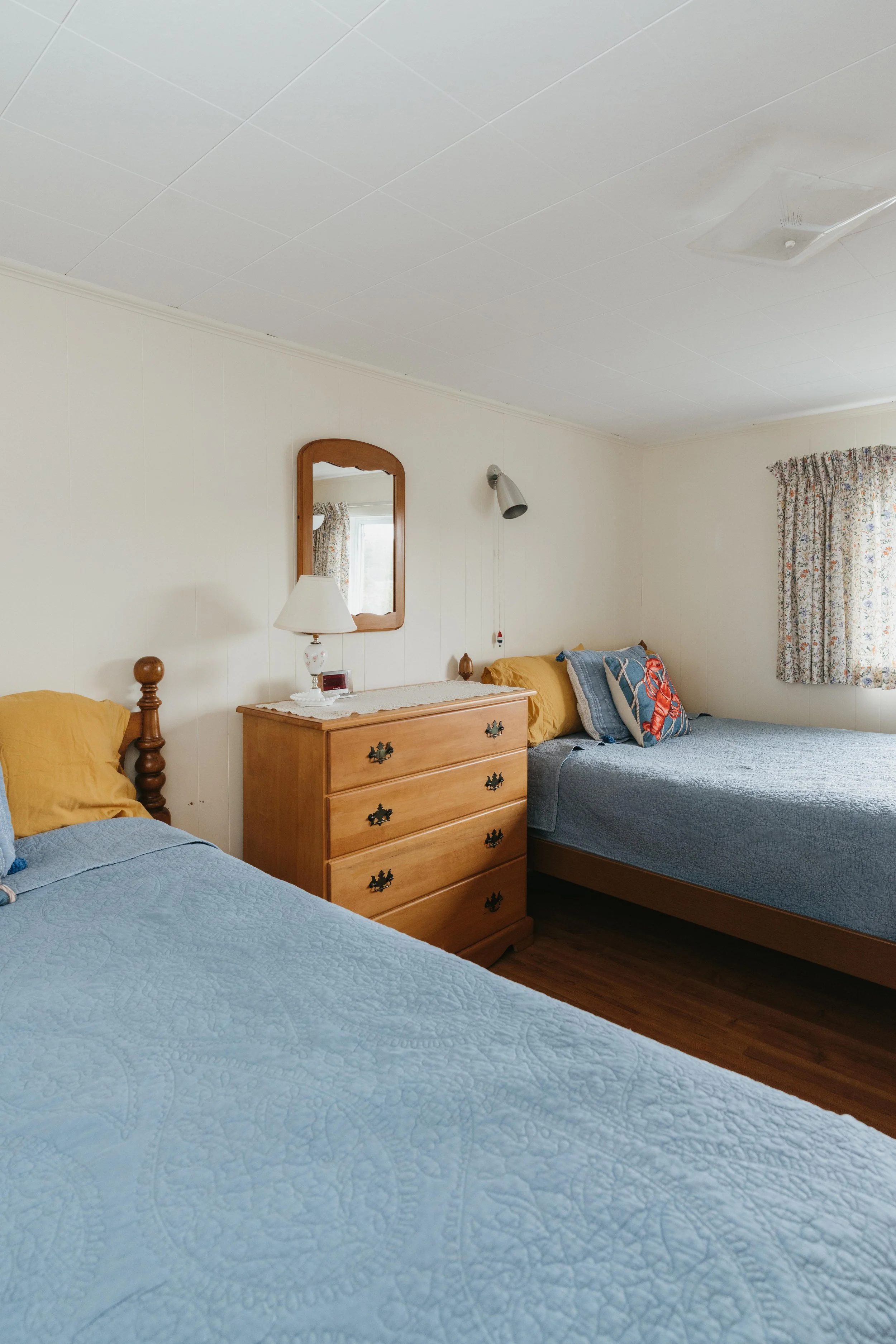 A bedroom with two beds, a wooden dresser with a lamp, mirror, and clock, and floral curtains.