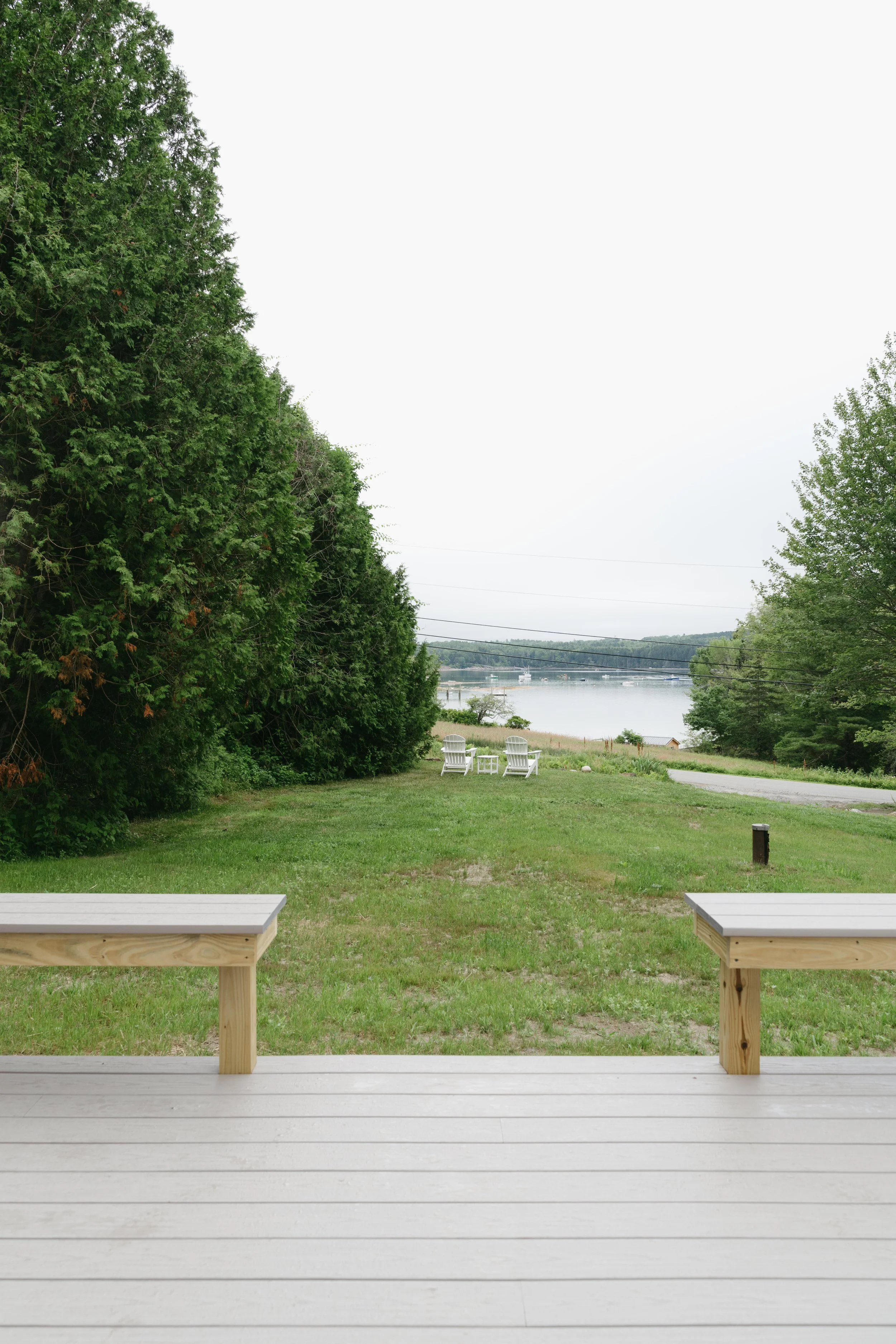 View of a backyard with a wooden deck, grass lawn, trees, and a view of a body of water with boats in the distance.