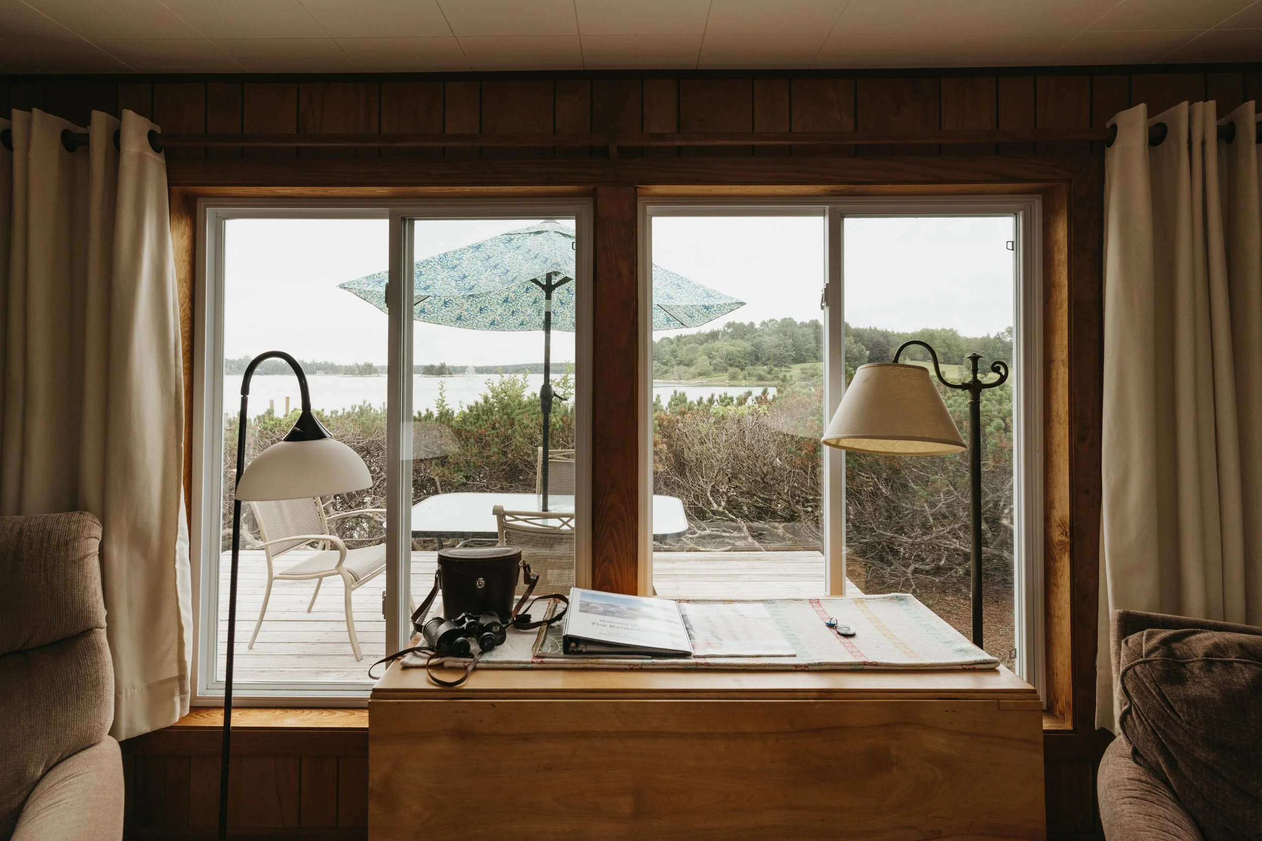 Interior view of a room looking out through large sliding glass doors to a deck with outdoor furniture, with an umbrella in the background, over a landscape with trees and water.