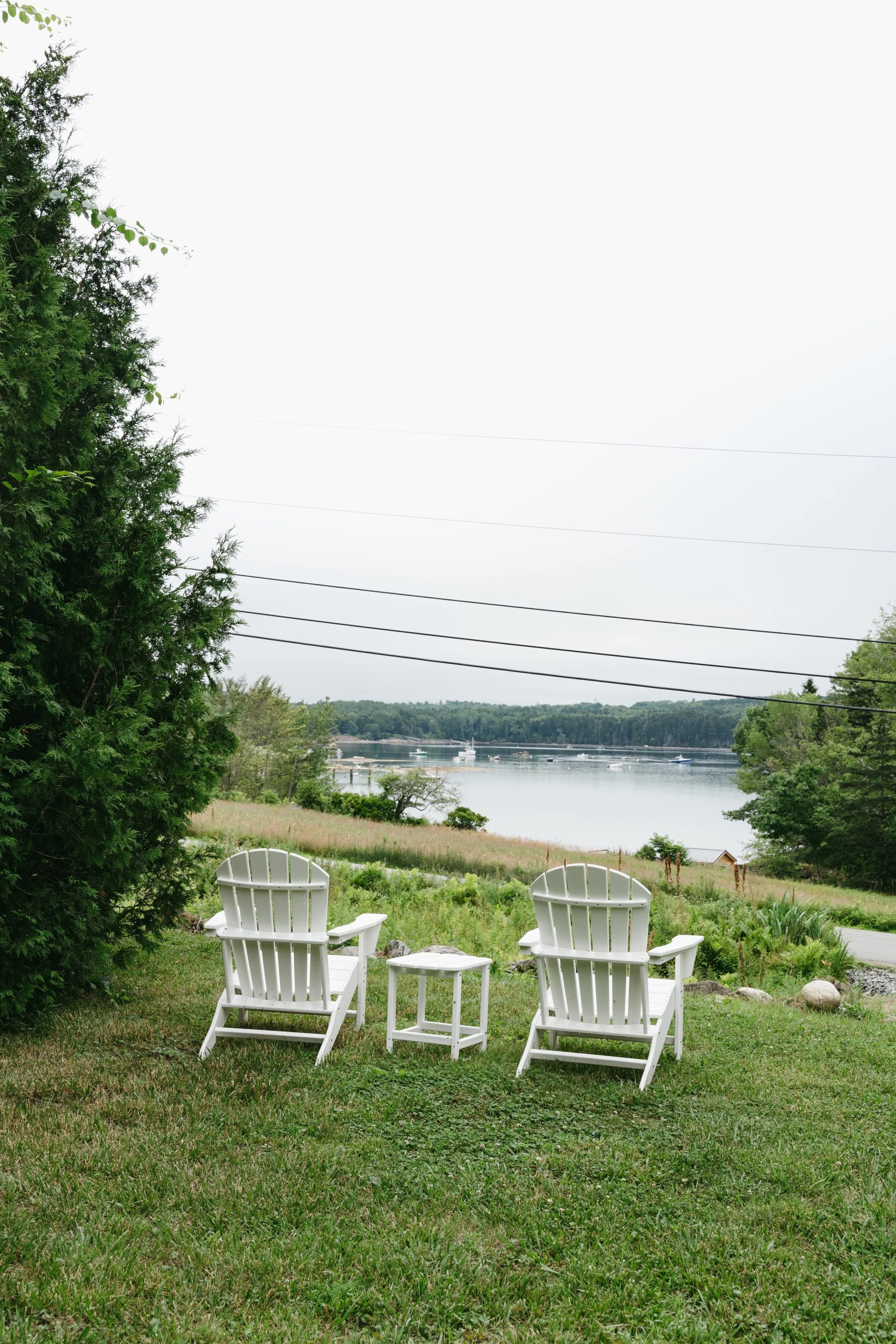 Two white Adirondack chairs and a small side table on a grassy lawn overlooking a body of water with trees and boats in the distance.