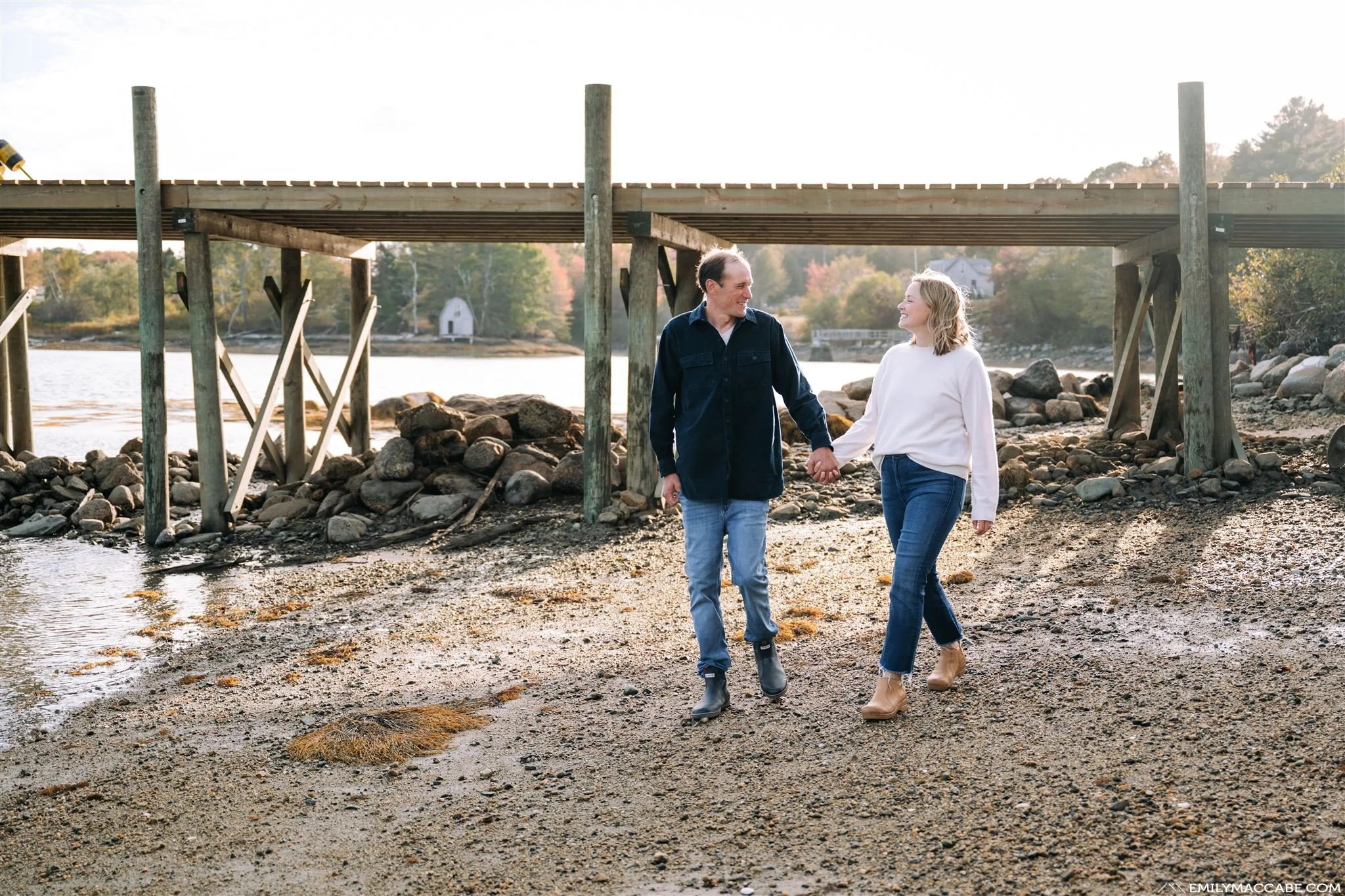A man and woman walking hand in hand along a rocky shoreline near a timber pier, with trees and water in the background.