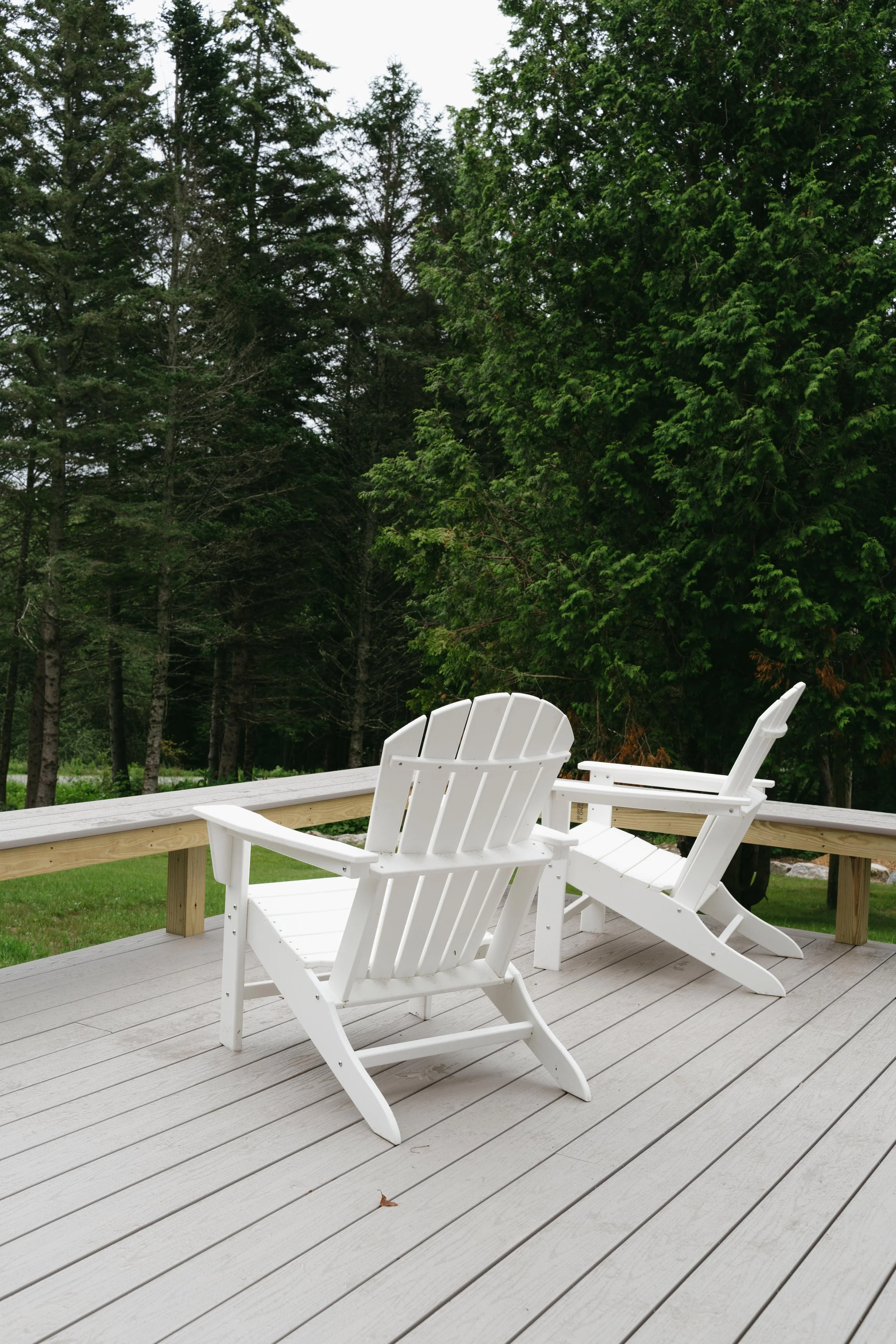White Adirondack chairs on a wooden deck overlooking a green yard with trees.