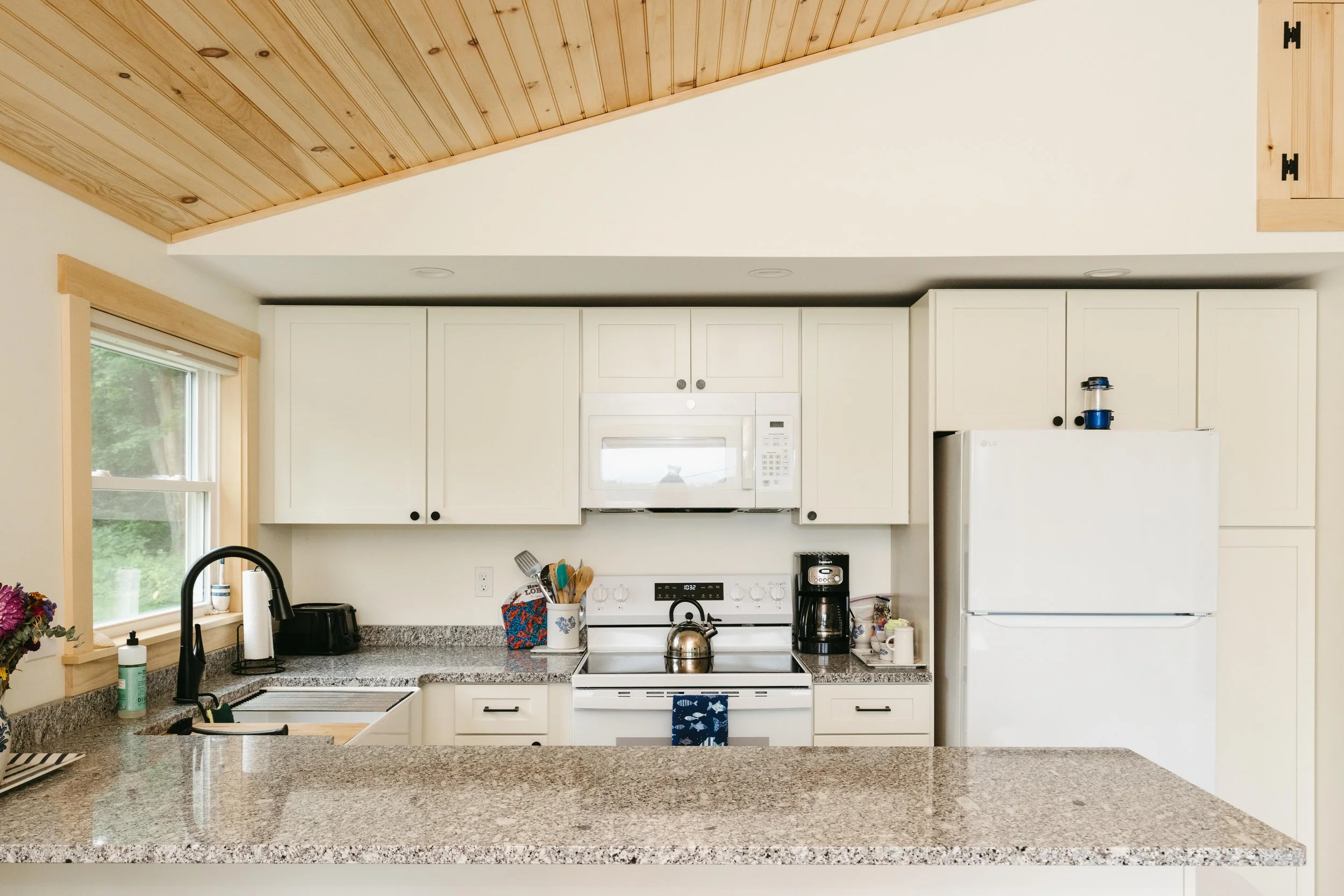 Kitchen with white cabinets, granite countertop, black faucet, stovetop with kettle, microwave, coffee maker, refrigerator, and window with greenery outside.
