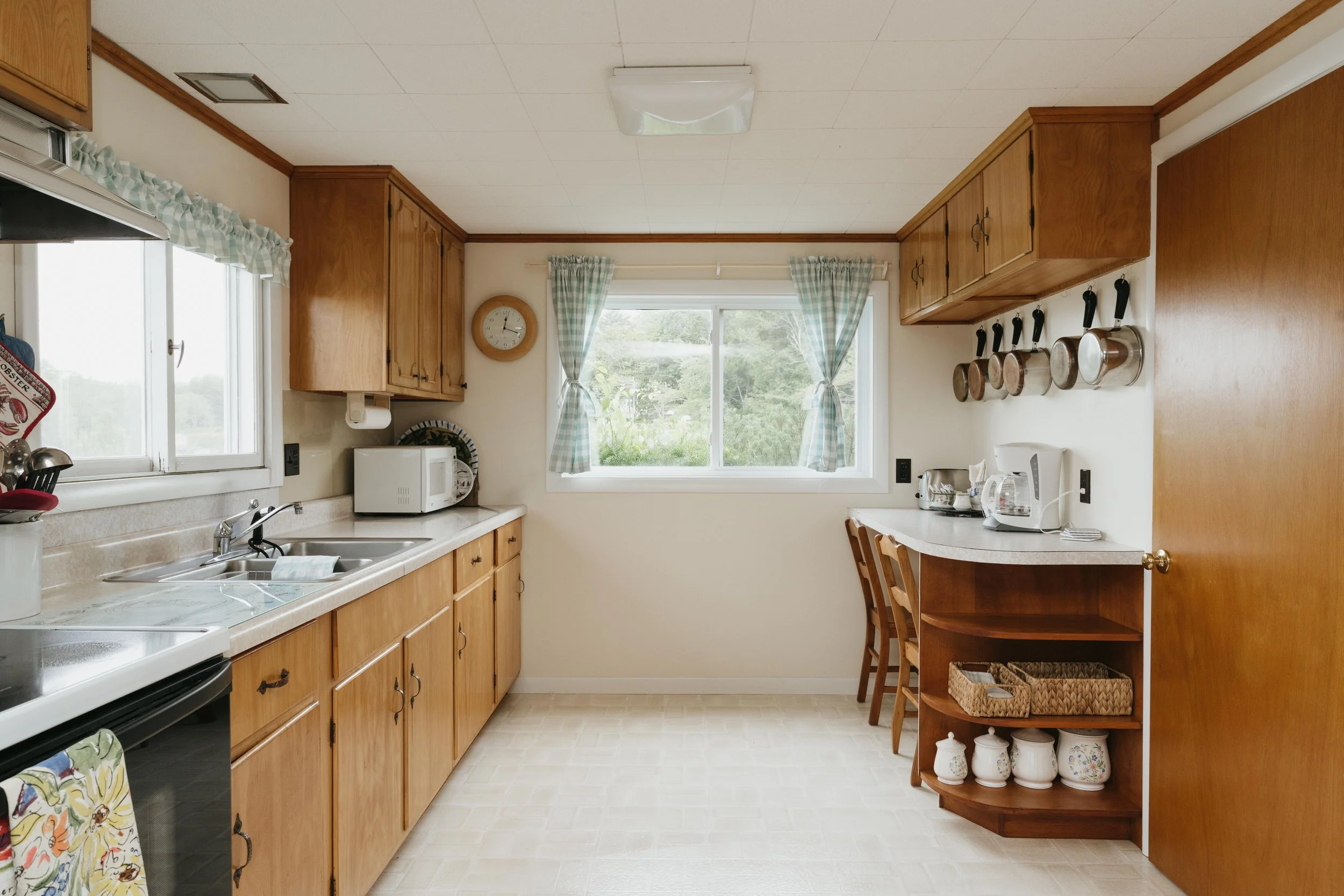 Kitchen with wooden cabinets, a window with checkered curtains, and various kitchen appliances including a microwave, toaster, and coffee maker. There are hanging pots and a clock on the wall.