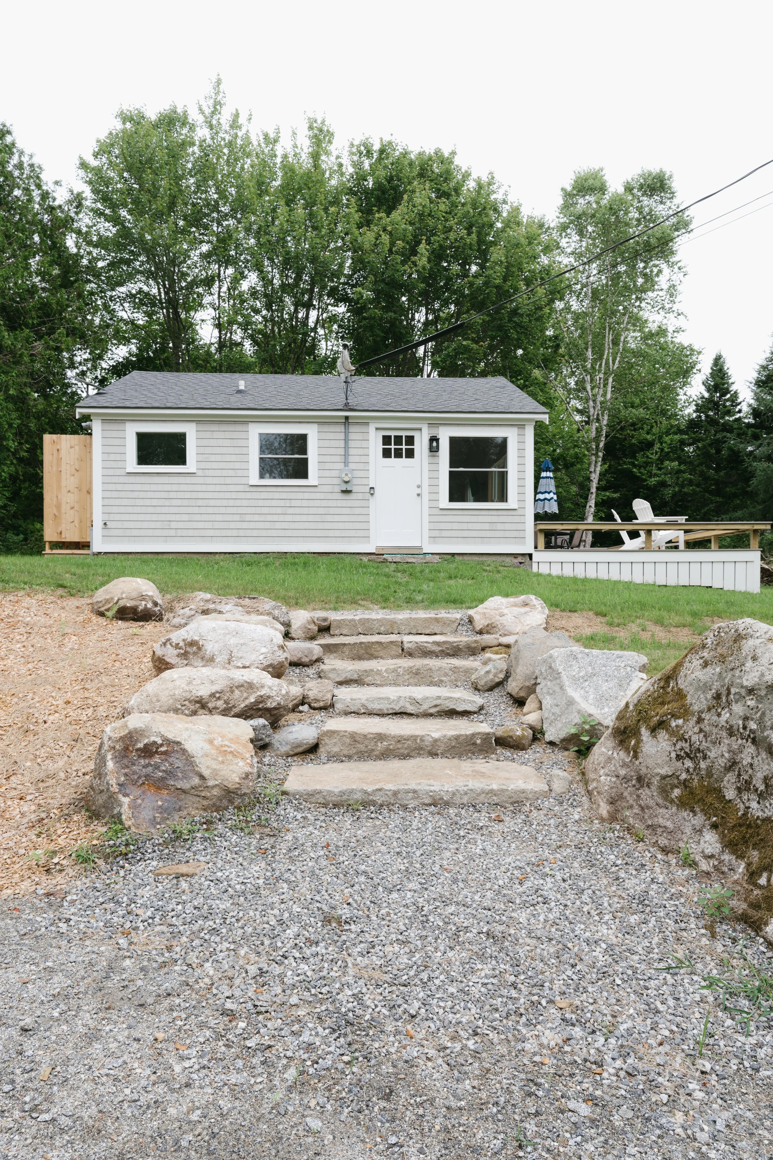 Small white house with a gray roof, surrounded by green trees, with stone steps and large rocks leading to the front door, and a gravel pathway in the front yard.