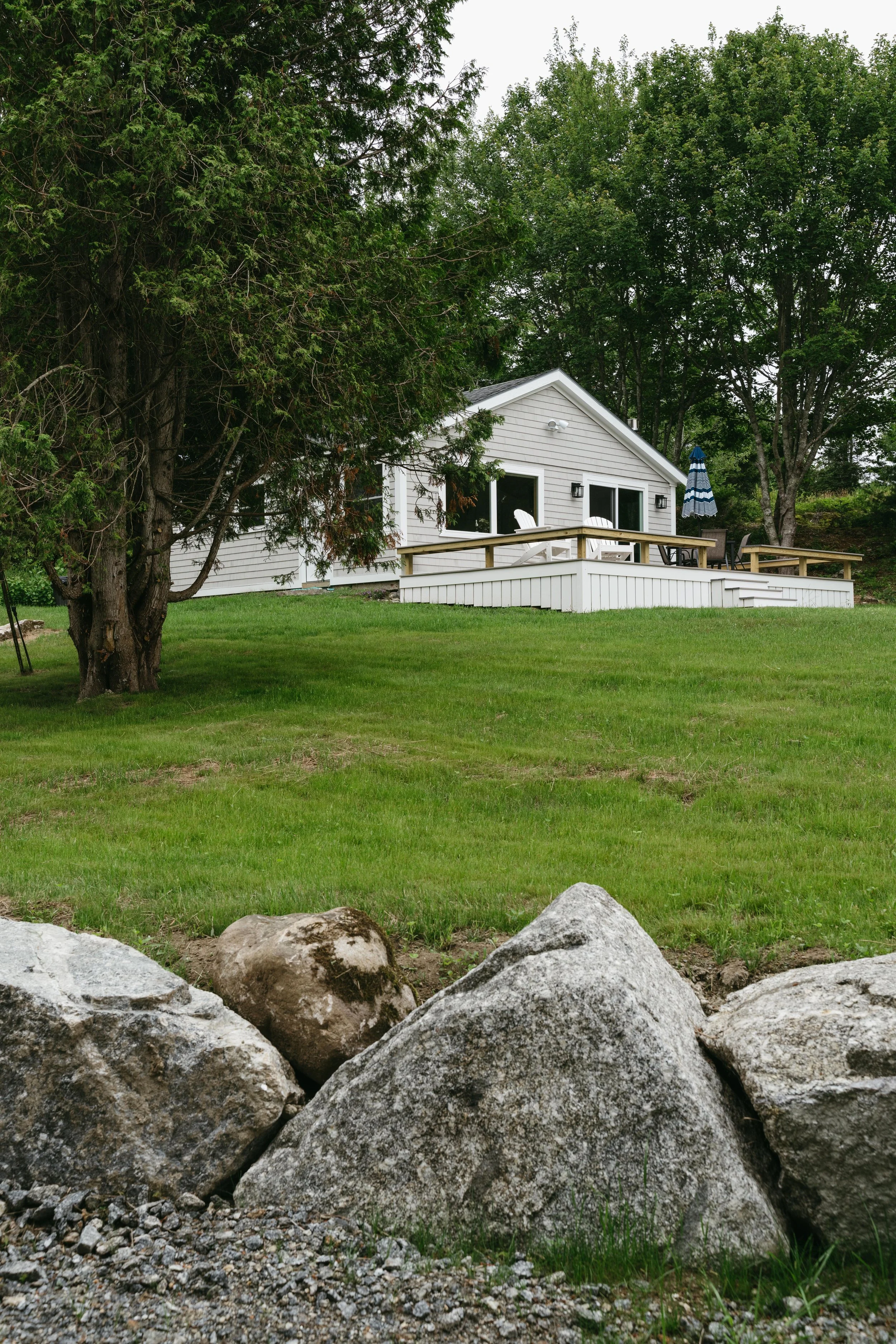 A house with a deck overlooking a lush green yard, trees in the background, and large rocks in the foreground.