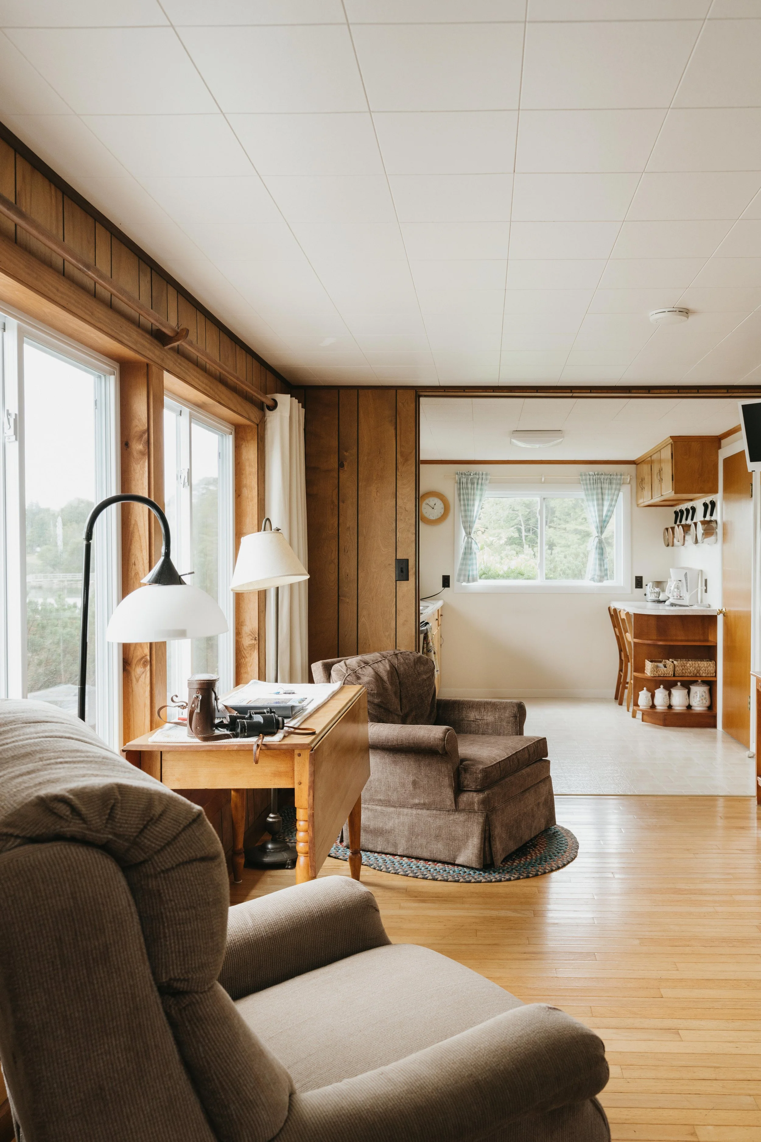 A cozy living room with wooden paneling, large windows with white curtains, and a sitting area with brown and beige armchairs. There is a wooden side table with a lamp, binoculars, and magazines on it. The kitchen area with a small counter, chairs, a