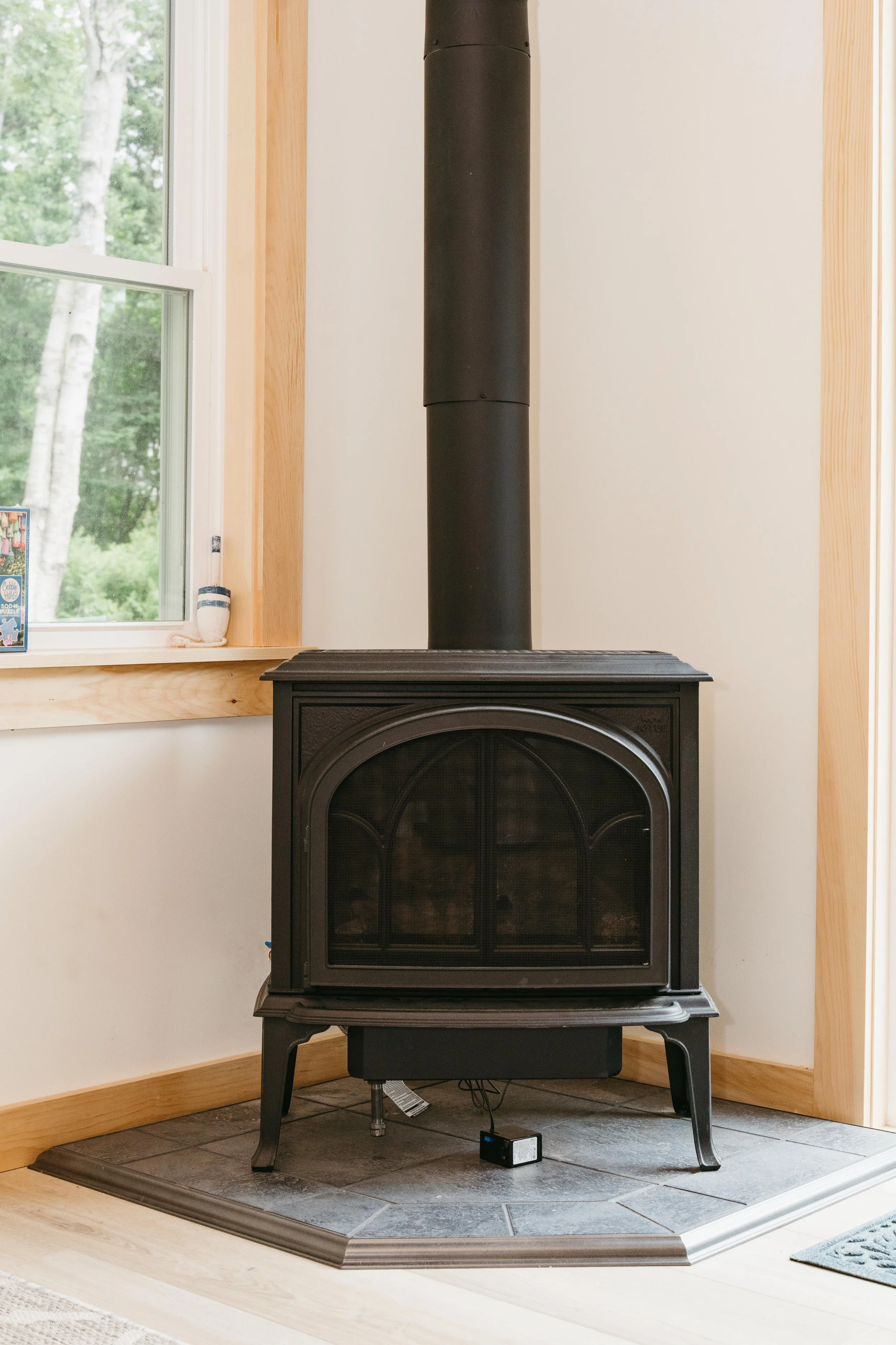 Black wood stove with a metal pipe venting through the ceiling, placed on a tiled hearth in a room with light walls and wooden trim. There is a window to the left showing green trees outside.