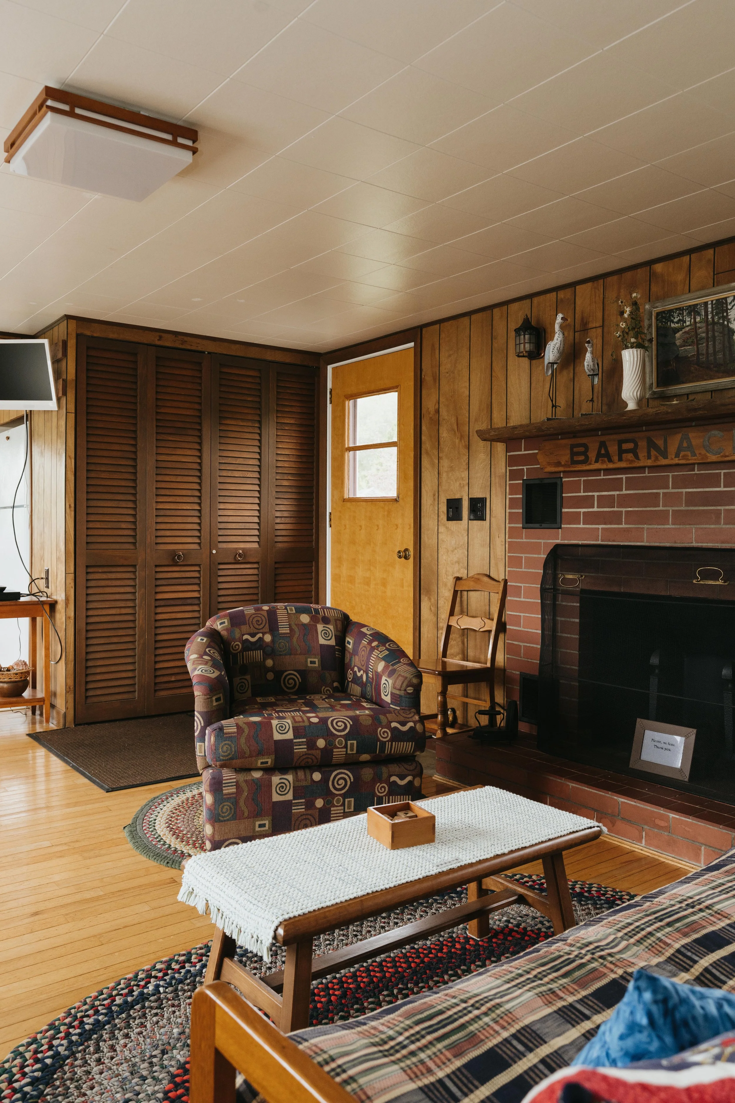 A cozy living room with wood-paneled walls, a brick fireplace, a colorful patterned armchair, a wooden coffee table with white cloth, plaid sofa, wooden chair, and closed brown louvered closet doors.