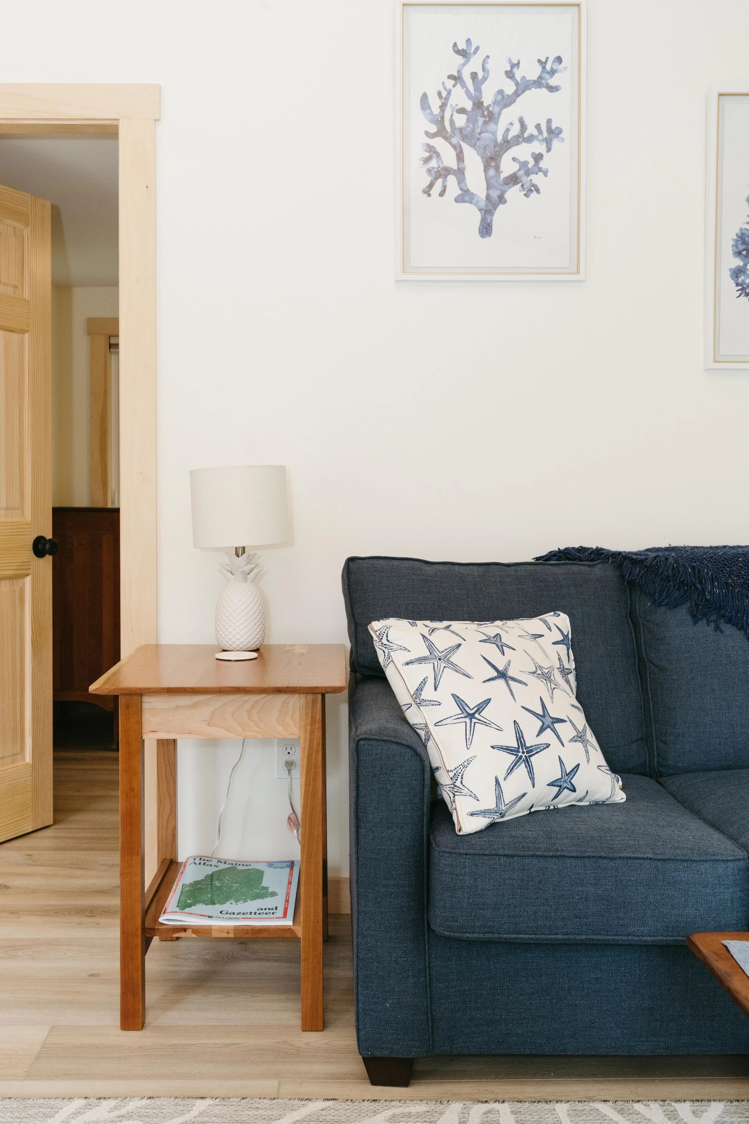 A cozy living room corner with a dark grey sofa, a pillow with starfish pattern, a wooden side table with a pineapple-shaped lamp, and framed underwater-themed artwork on the white wall.