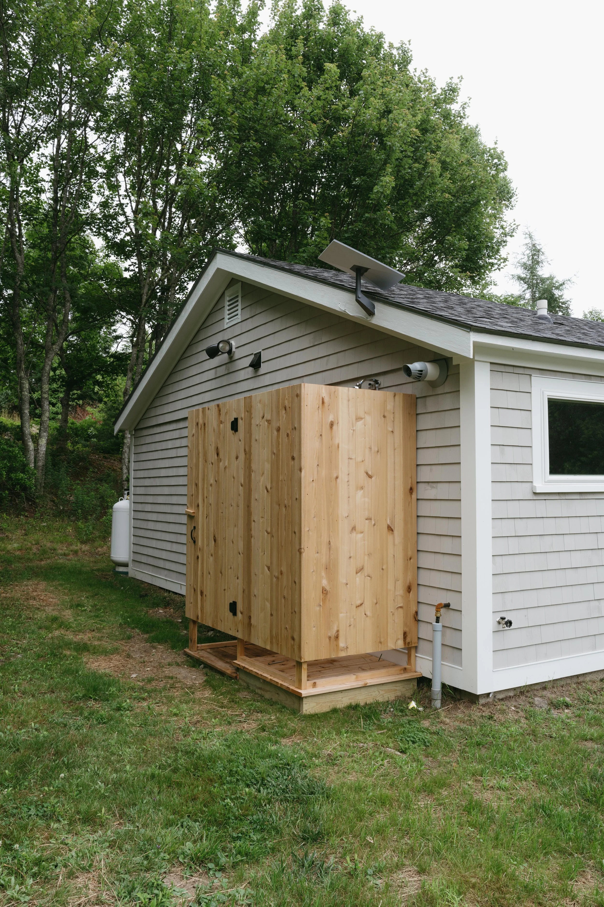 Exterior of a house with a wooden outdoor shower setup attached to the side, surrounded by green trees and grass.