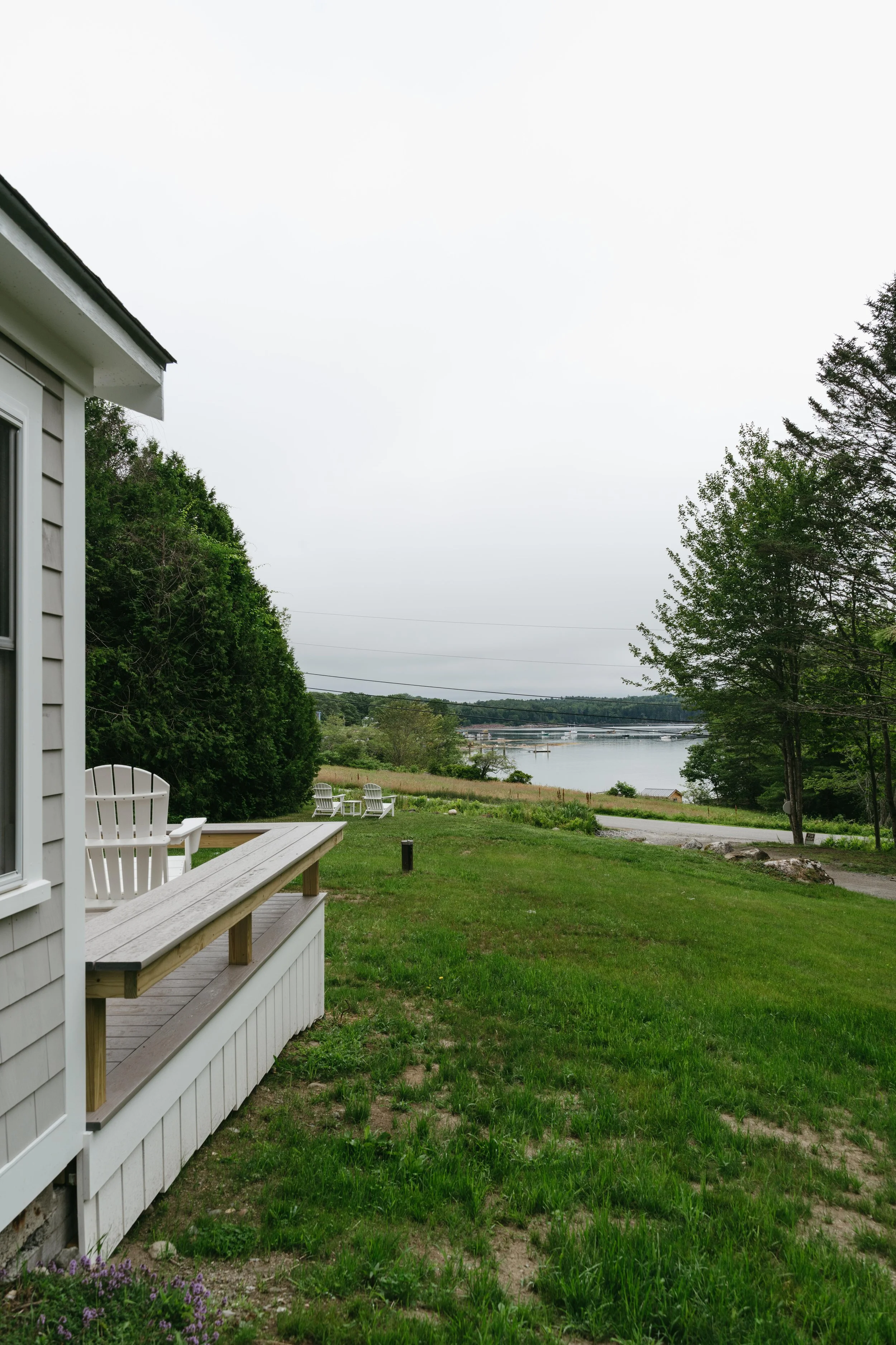 A backyard scene with a white house on the left, outdoor white chairs, a small deck, green grass, trees, and a body of water in the background under an overcast sky.