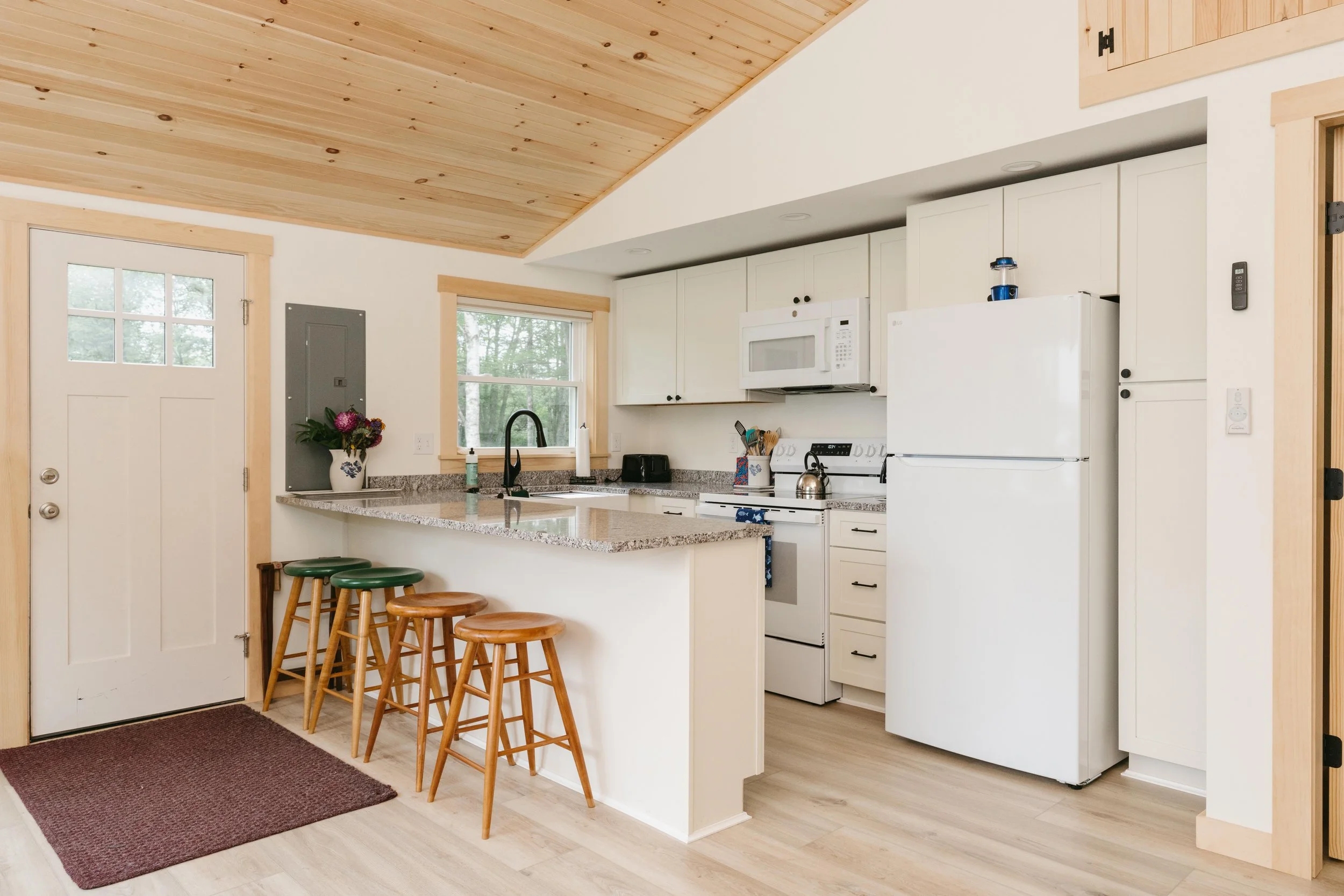 Kitchen with white cabinets, a granite countertop island with three wooden and green stools, a white refrigerator, microwave, stove, and window with trees outside.