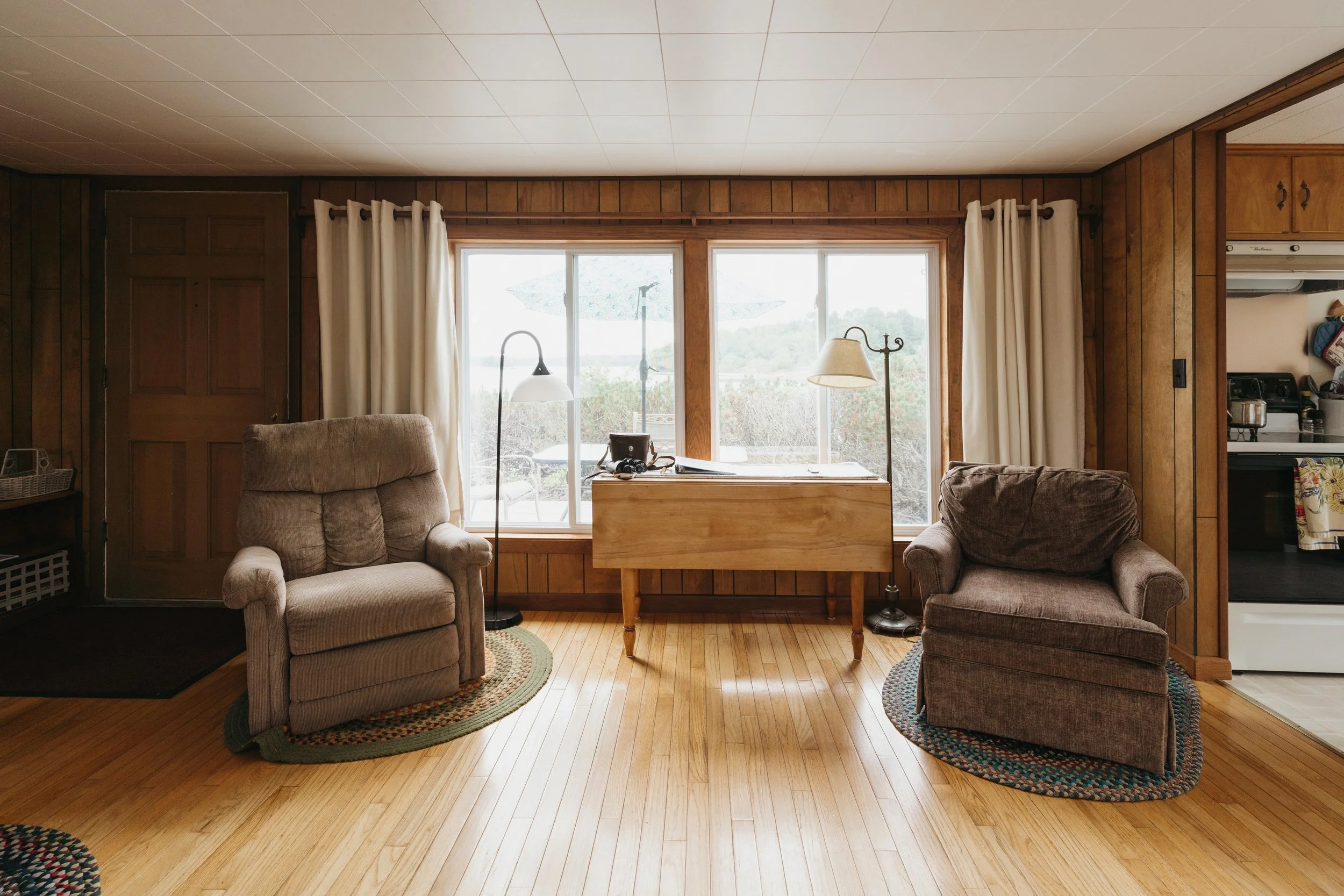 A living room with wood-paneled walls, a large window with curtains, two armchairs on oval rugs, a small wooden table, and a sliding door leading outside.