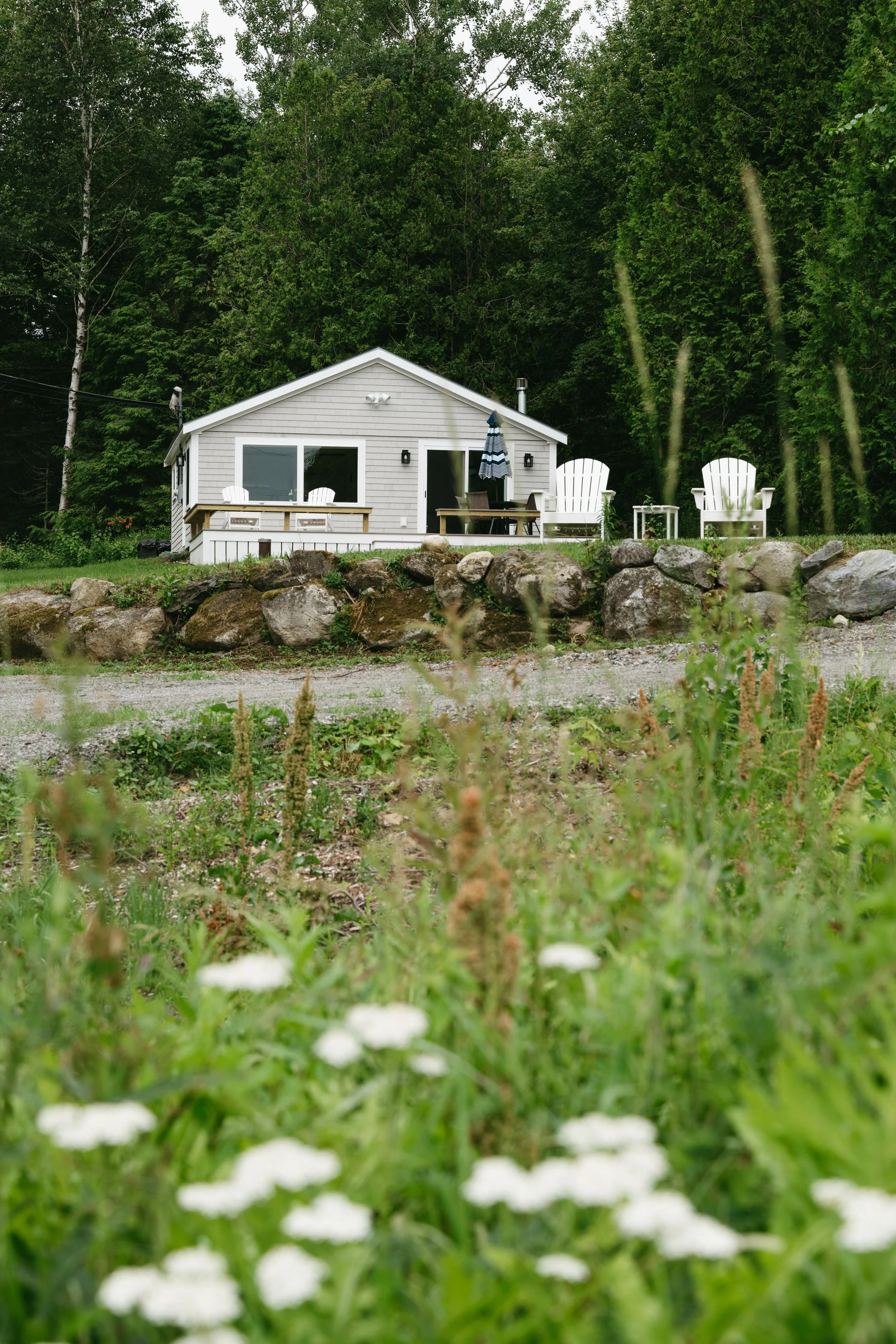 A small house with a deck, surrounded by trees, with two large white chairs and an umbrella outside, in a natural, rural setting.