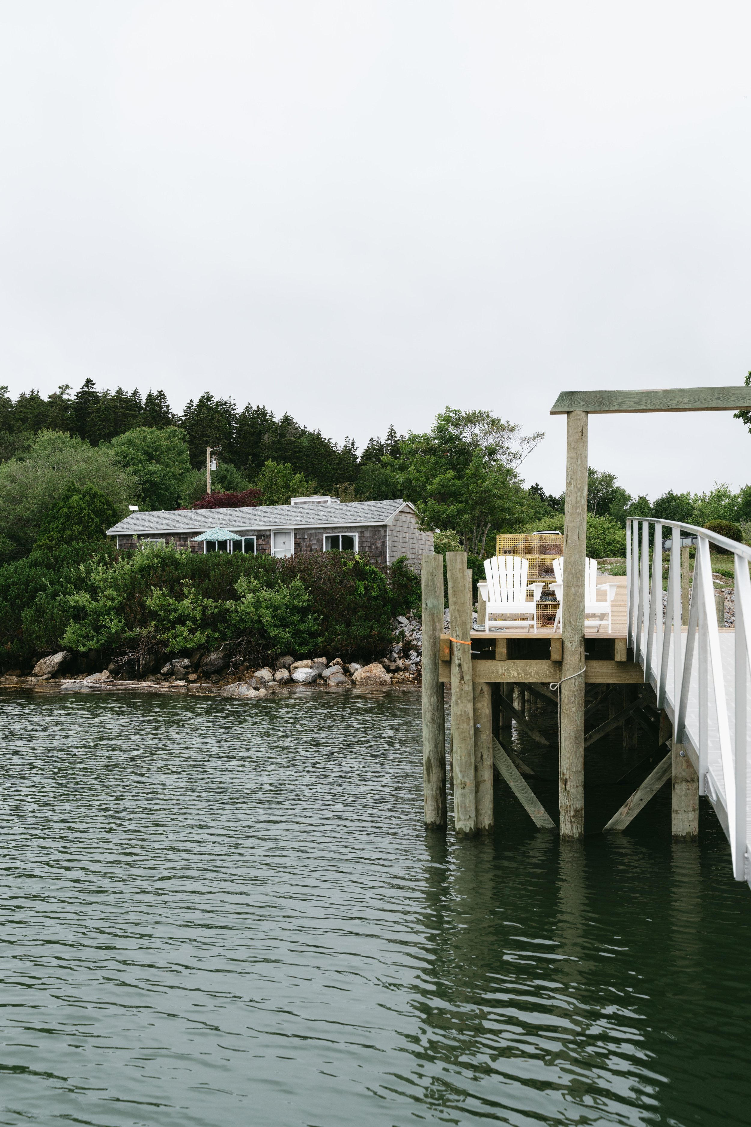 View of a waterfront house with a dock and white chairs on the deck, surrounded by trees and rocks on a cloudy day.