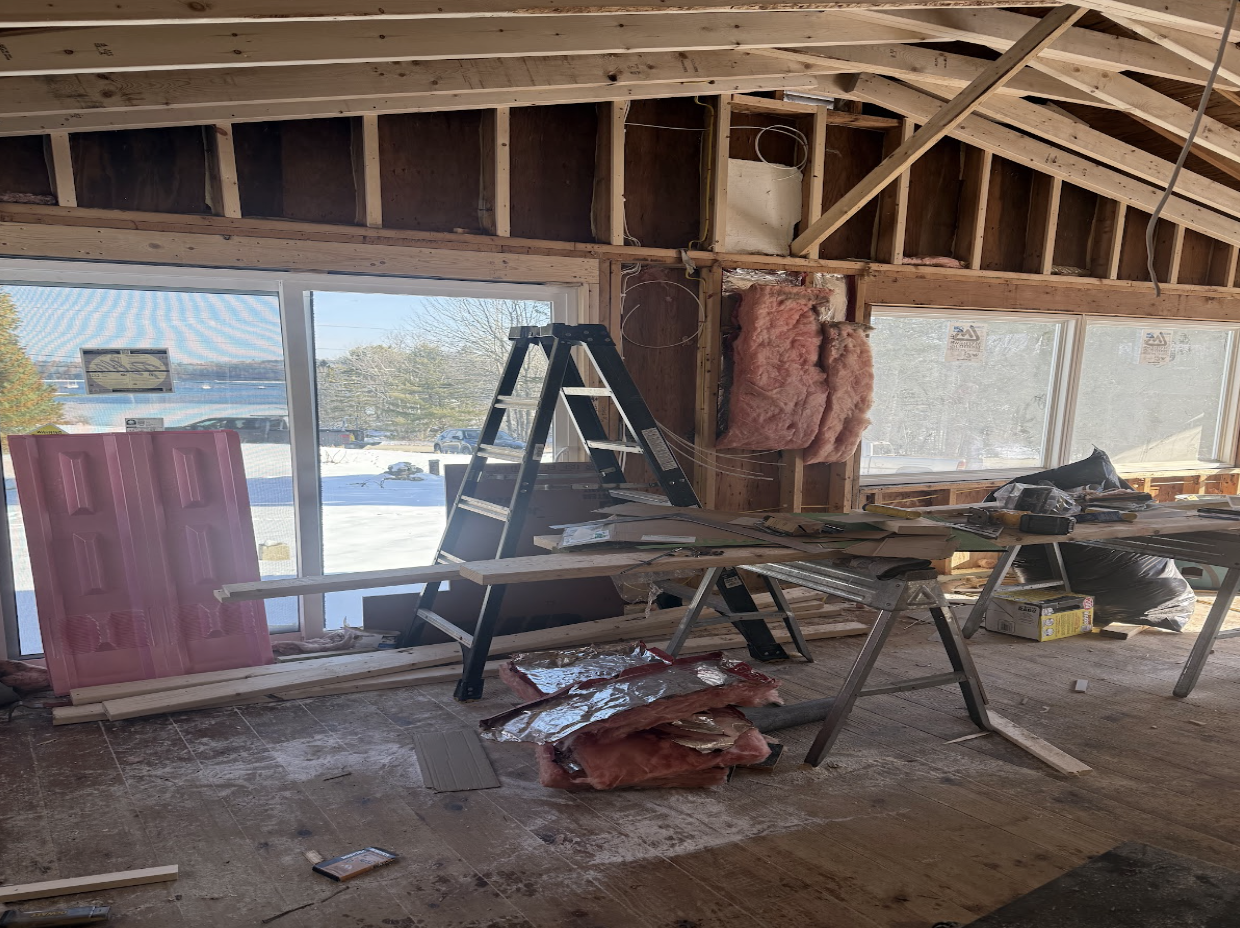 Interior of a house under construction with exposed wooden framing, insulation, a ladder, and construction tools, with large windows showing a snowy landscape outside.