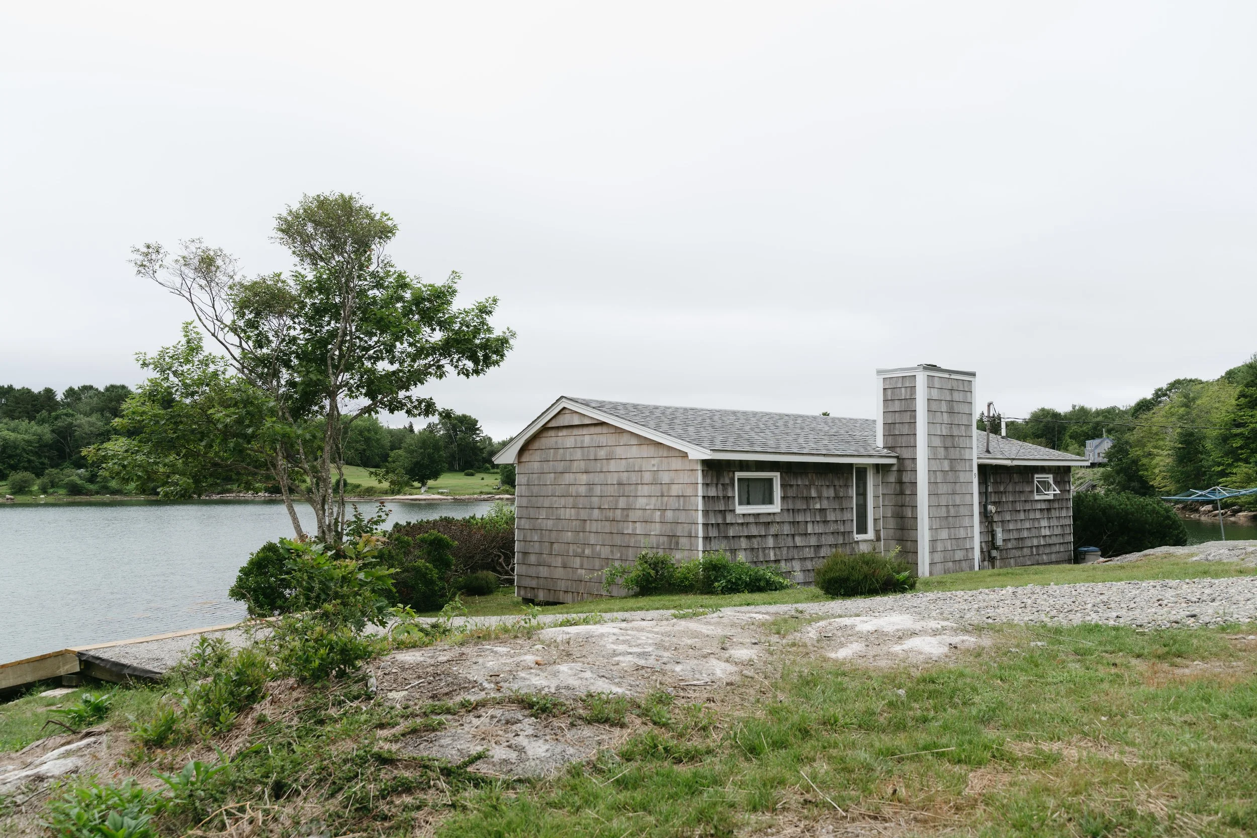 A small house with gray weathered wooden siding and a gray shingled roof by a lake, with a tree and shrubs in the yard.