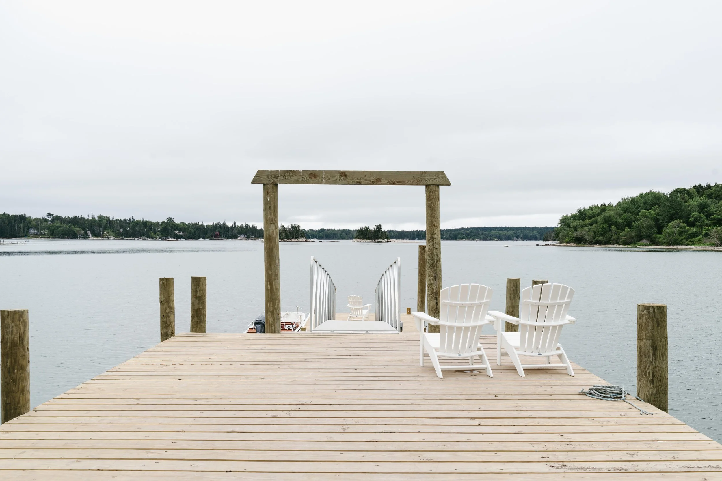 A wooden dock extending into a calm lake, with two white Adirondack chairs on the right, and a simple wooden pergola frame at the end of the dock. The scene is overcast, with trees lining the distant shoreline.