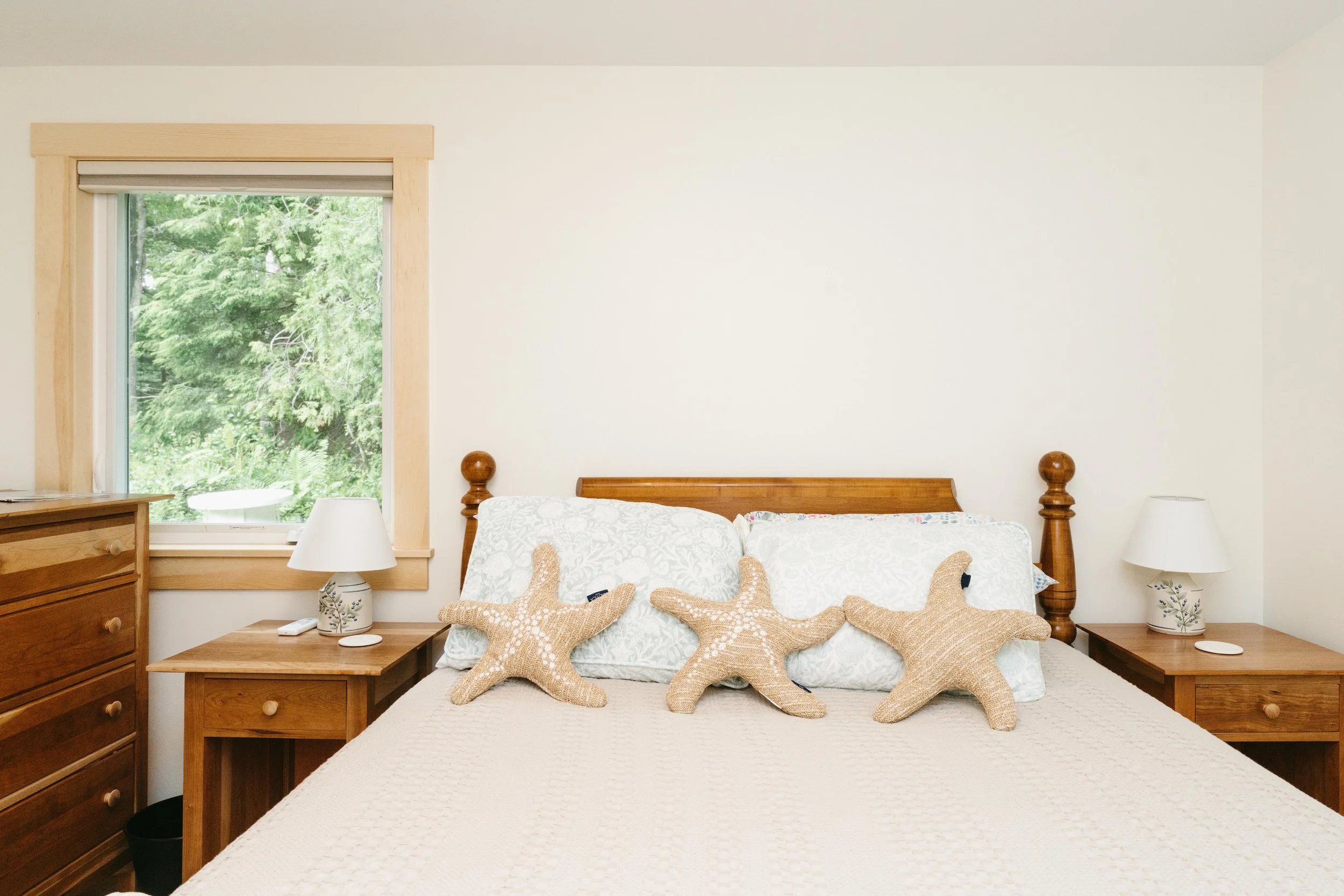 A bedroom with a wooden bed frame, white bedding, and three starfish pillows, flanked by wooden nightstands with white lamps and a window showing green trees outside.